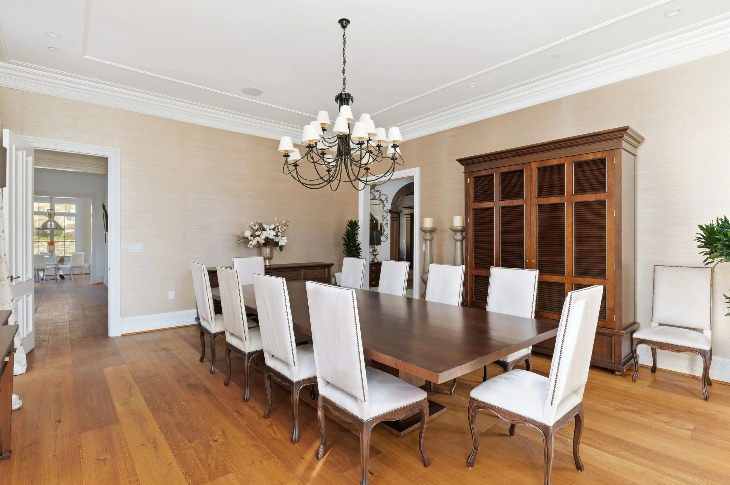 A dining room with a large wooden table surrounded by white upholstered chairs. A black metal chandelier with white lampshades hangs above the table. There's a wooden cabinet with louvered doors on the wall, a vase of flowers on a sideboard, and some