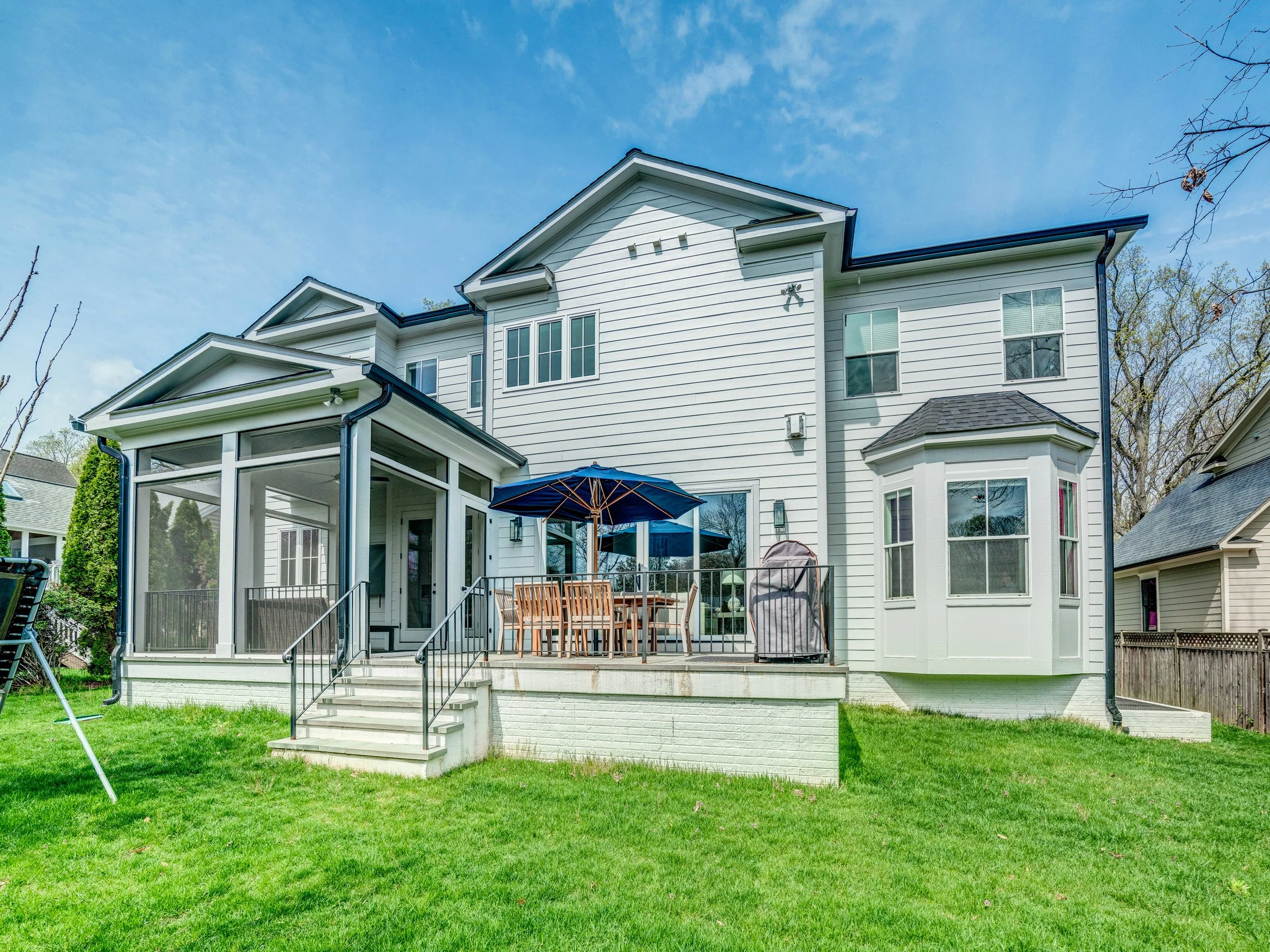 Backyard view of a white two-story house with a screened porch and a patio set with an umbrella, surrounded by green grass and trees under a clear blue sky.