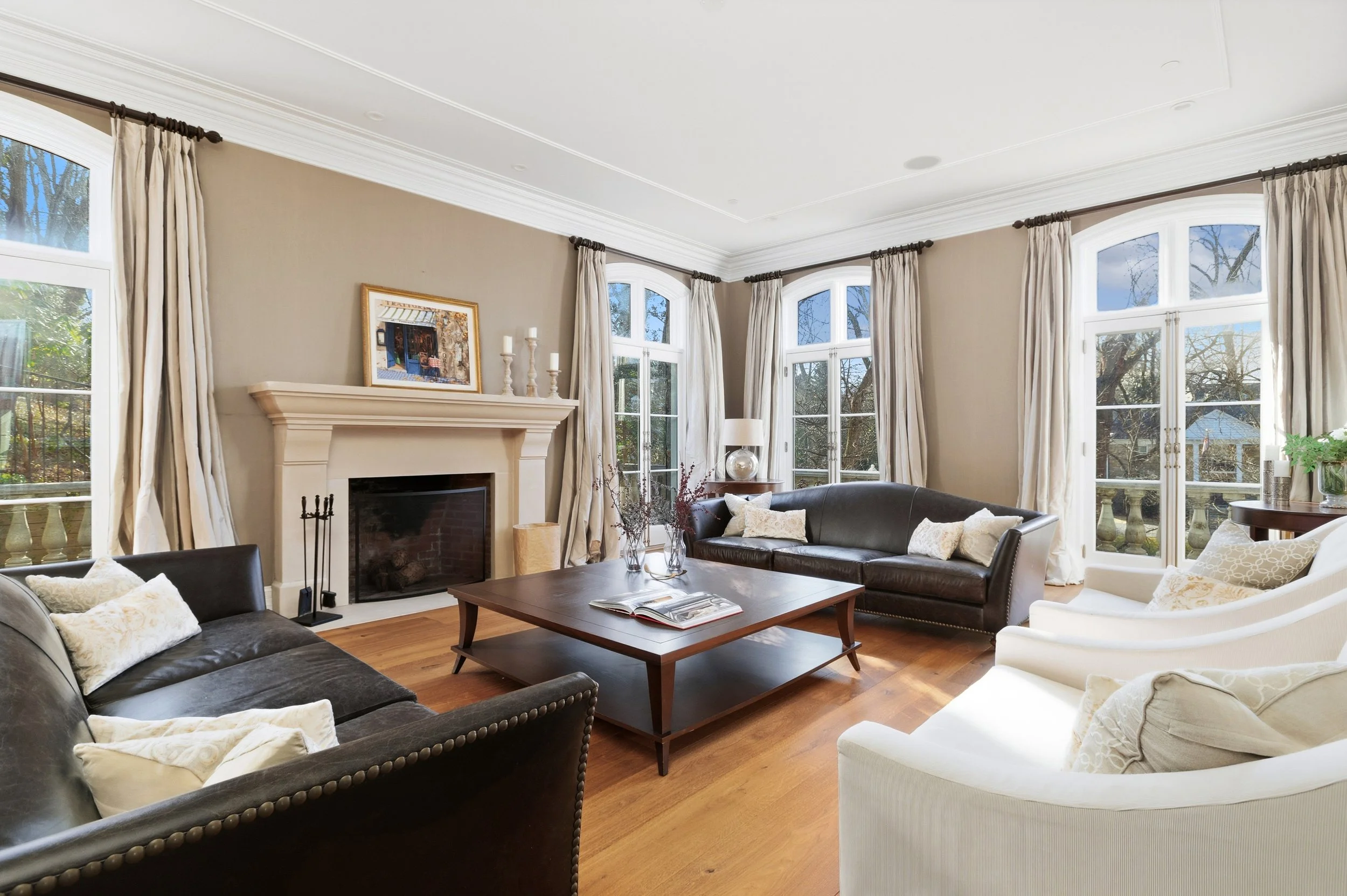 Living room with beige walls, white crown molding, large windows with white frames and cream curtains, a fireplace with artwork on the mantle, various sofas with cushions, a wooden coffee table with a book, a table lamp, and decorative candles.