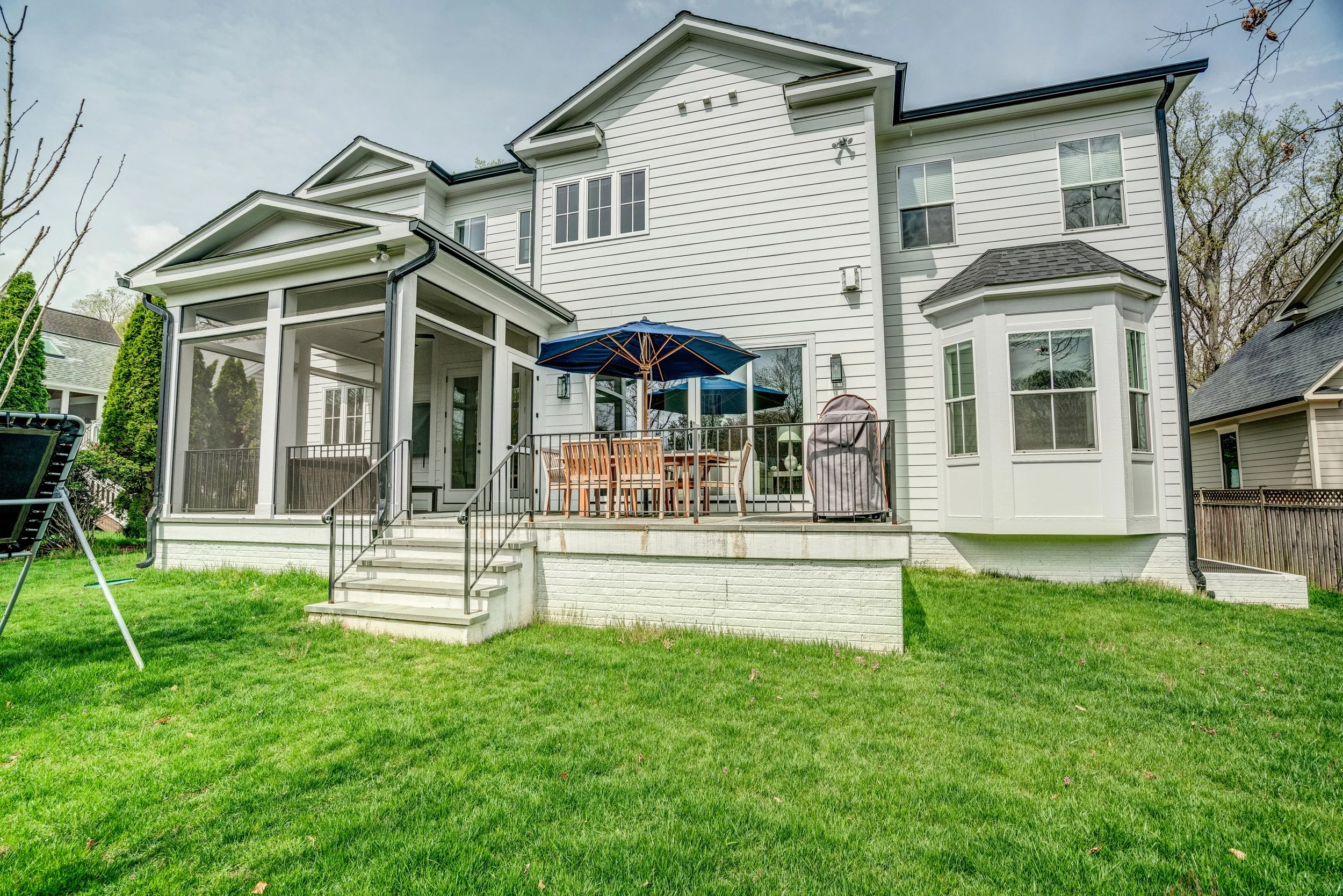Backyard view of a white, multi-story house with a screened porch, outdoor dining table with chairs, umbrella, and grass yard, surrounded by trees and neighboring houses.