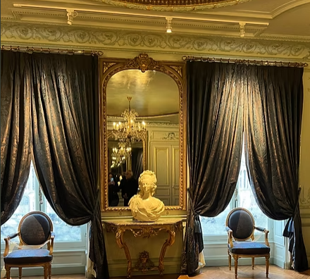 Elegant room with large window curtains, a framed mirror, a bust sculpture on a console table, and two chairs.