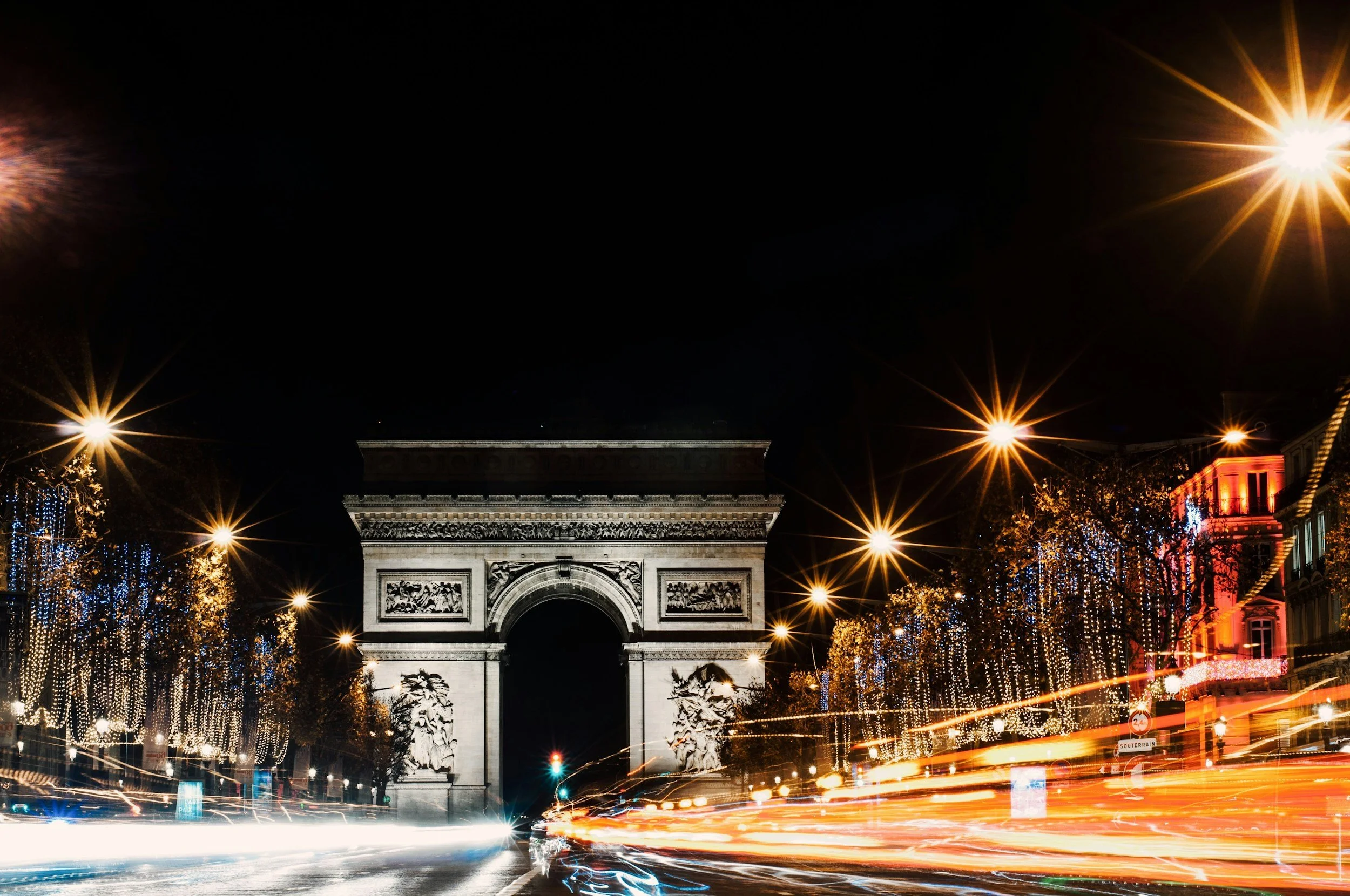 Nighttime view of the Arc de Triomphe in Paris, with festive lights and decorations on trees and buildings, and light trails from passing cars.