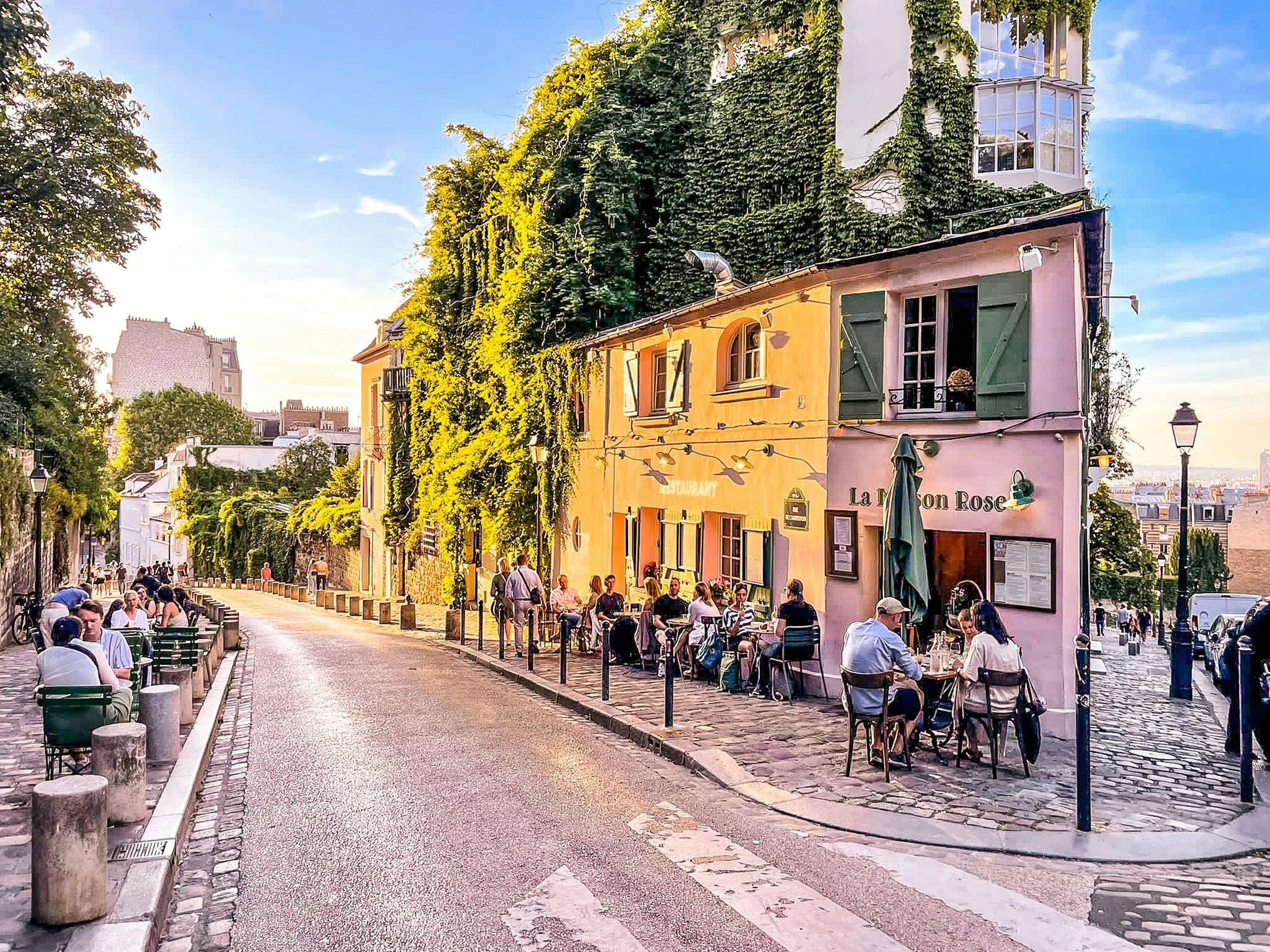 Outdoor cafe with people sitting at tables along a cobblestone street, colorful buildings covered with green ivy, and a clear blue sky.