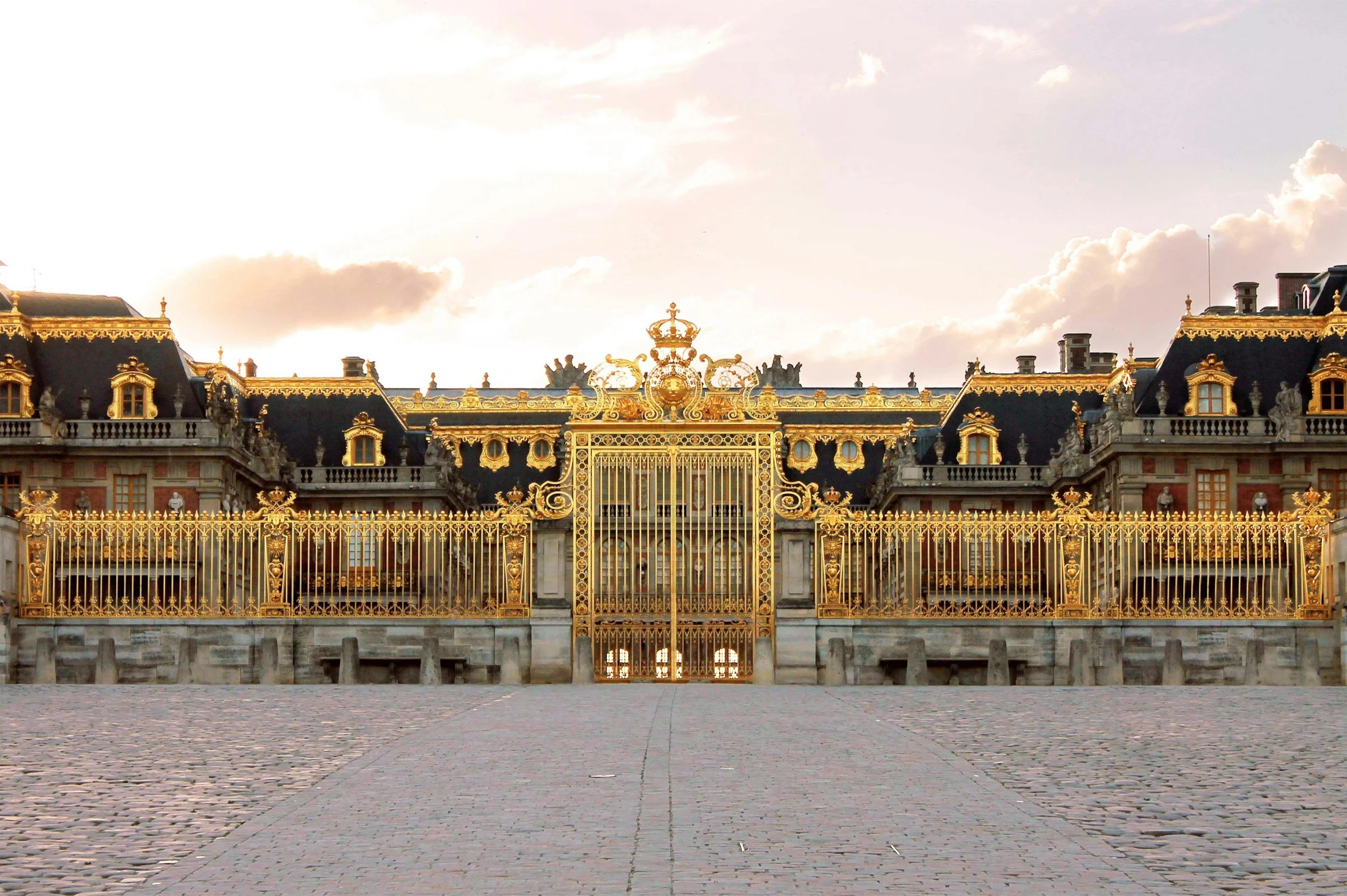 The image shows the ornate top of a historic building with intricate gold detailing, grand gates, and sculptures, set against a cloudy sky at sunset.