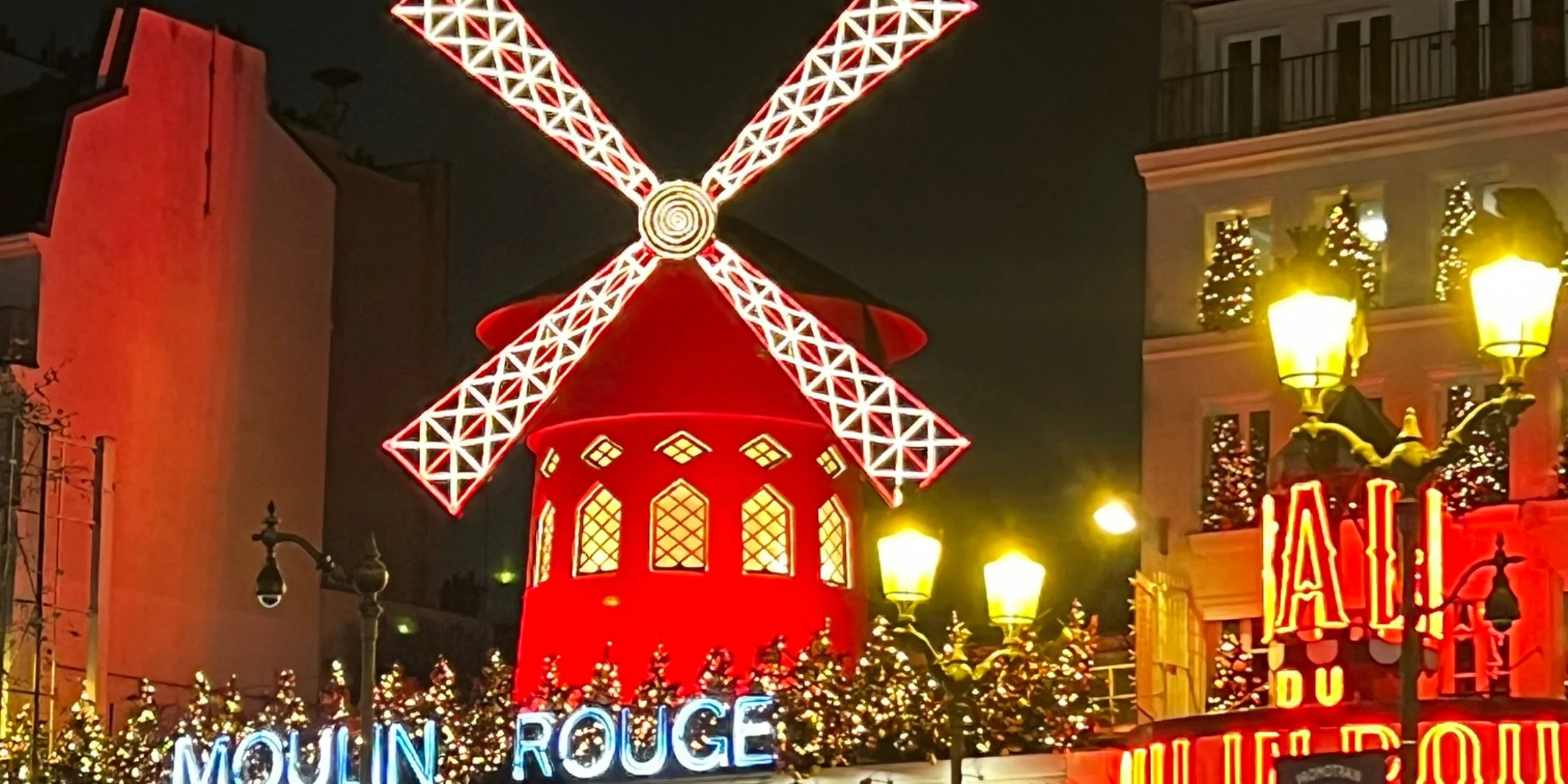 Decorative red windmill with illuminated blades at night, surrounded by streetlights and festive holiday decorations in a city street.