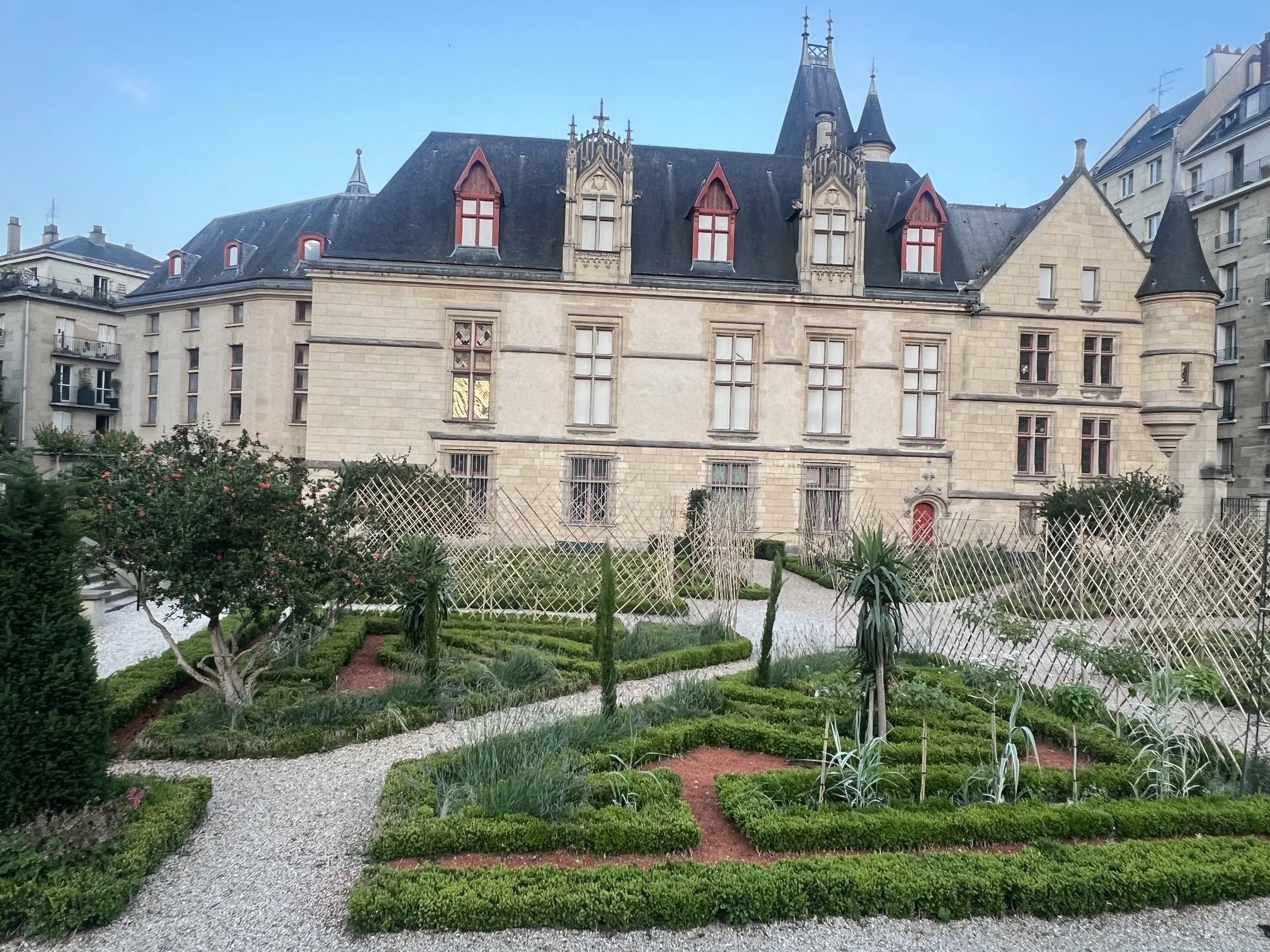 A historic building with gothic architectural features, surrounded by a neatly maintained garden with winding gravel pathways, shrubs, and young trees.
