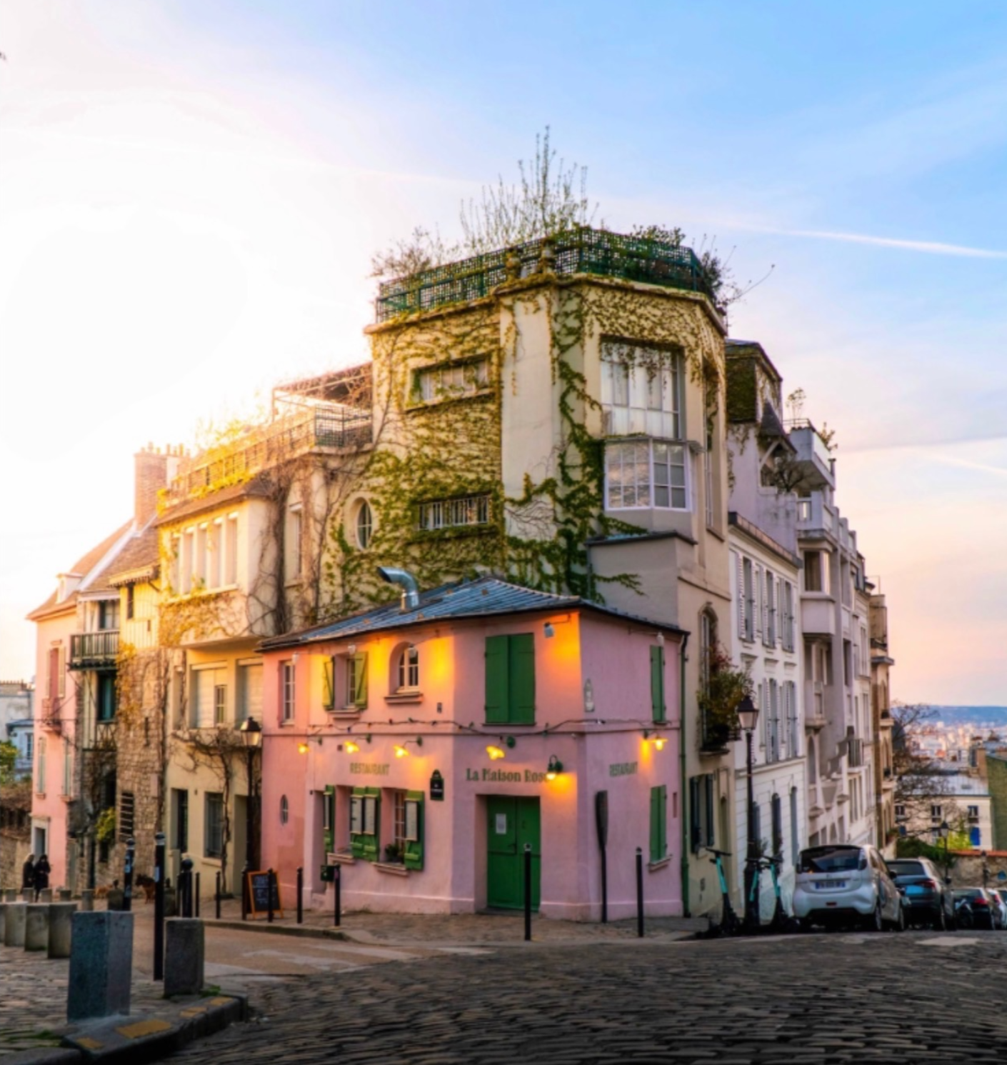 Colorful Parisian building with ivy-covered walls, pink storefront, and green shutters, on a cobblestone street at sunset.