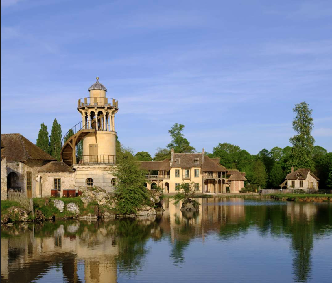 A picturesque scene of a historic European-style village with a water tower, surrounded by trees, houses, and a calm body of water reflecting the buildings and sky.