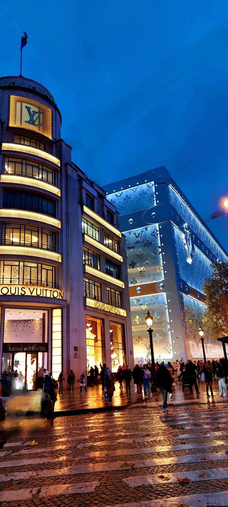 Nighttime street scene featuring a Louis Vuitton store, illuminated signage, and a large modern building with Louis Vuitton logos and designs, with people walking on wet cobblestone pavement.