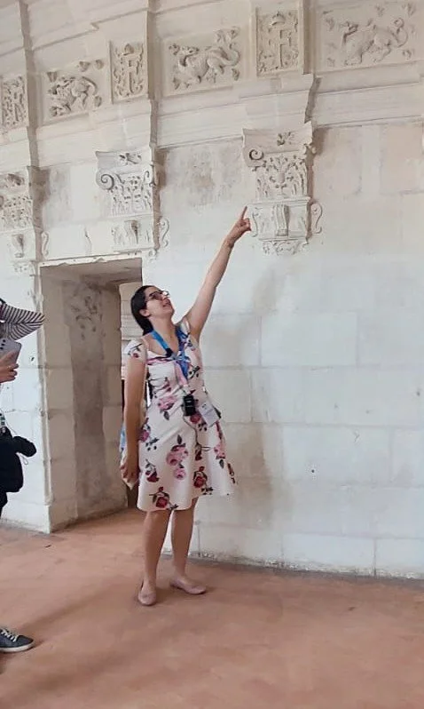 A woman in a floral dress pointing up at intricate stone carvings on an ancient white stone wall in a historical site.