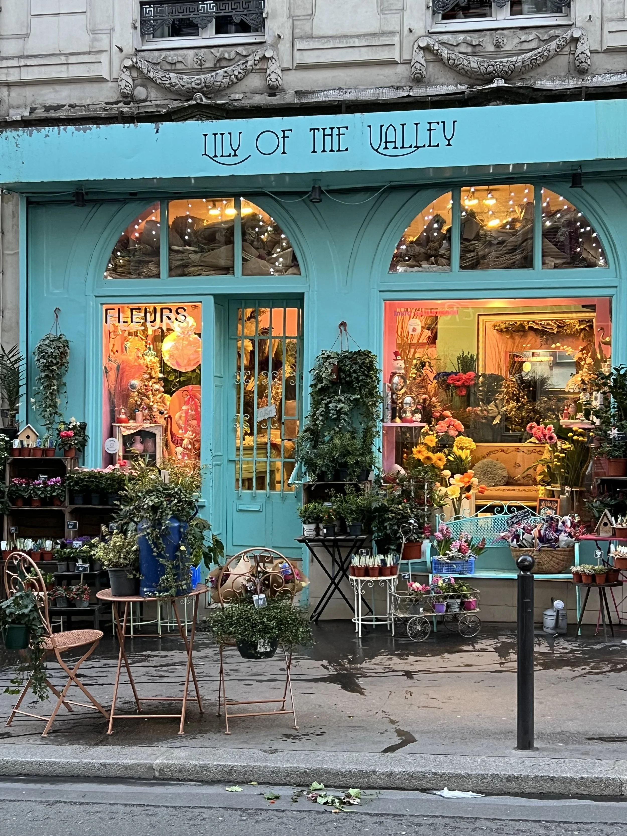 Flower shop with turquoise storefront, displaying various colorful flowers and plants outside, with chairs and tables on the street in front.