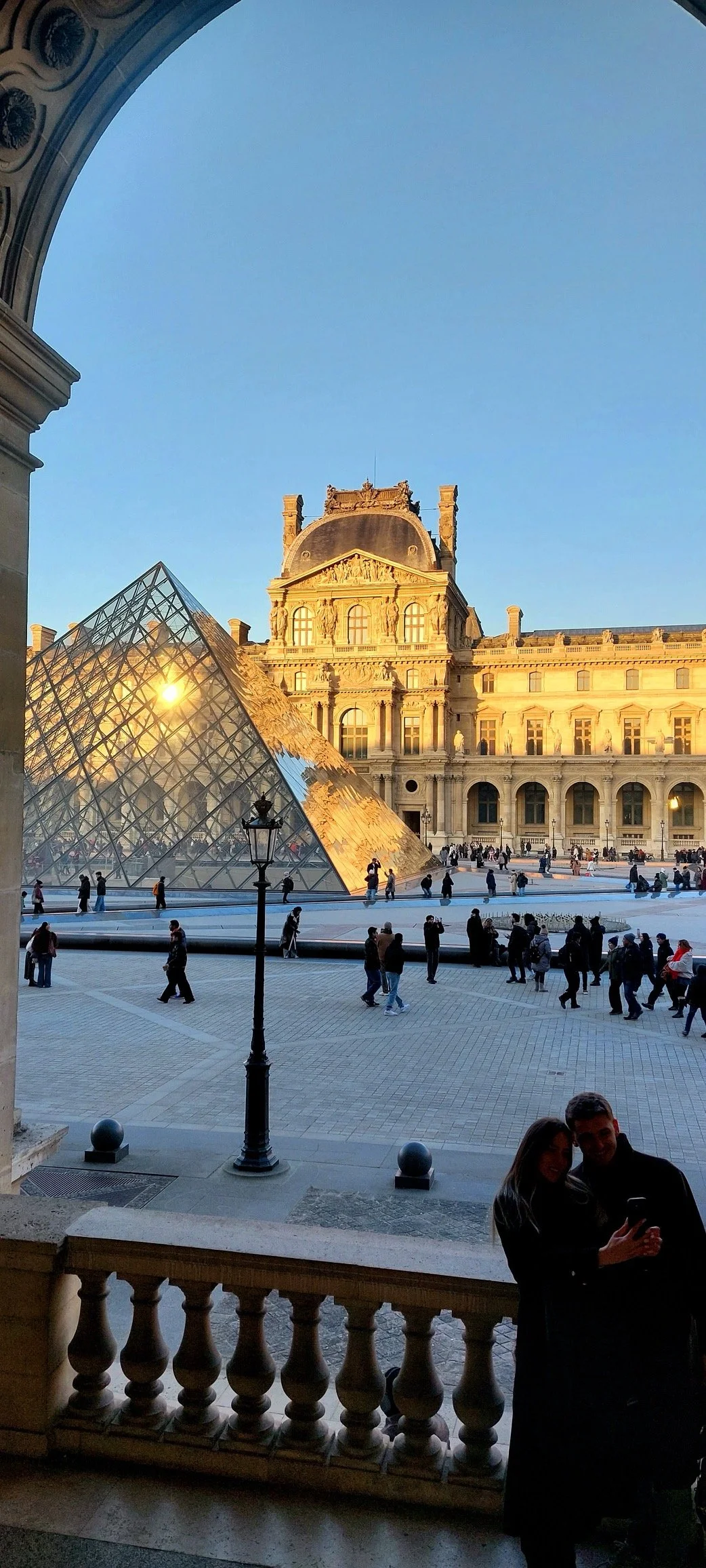 View of the Louvre Museum with its glass pyramid, classical building facades, and a crowd of tourists at sunset in Paris, France.