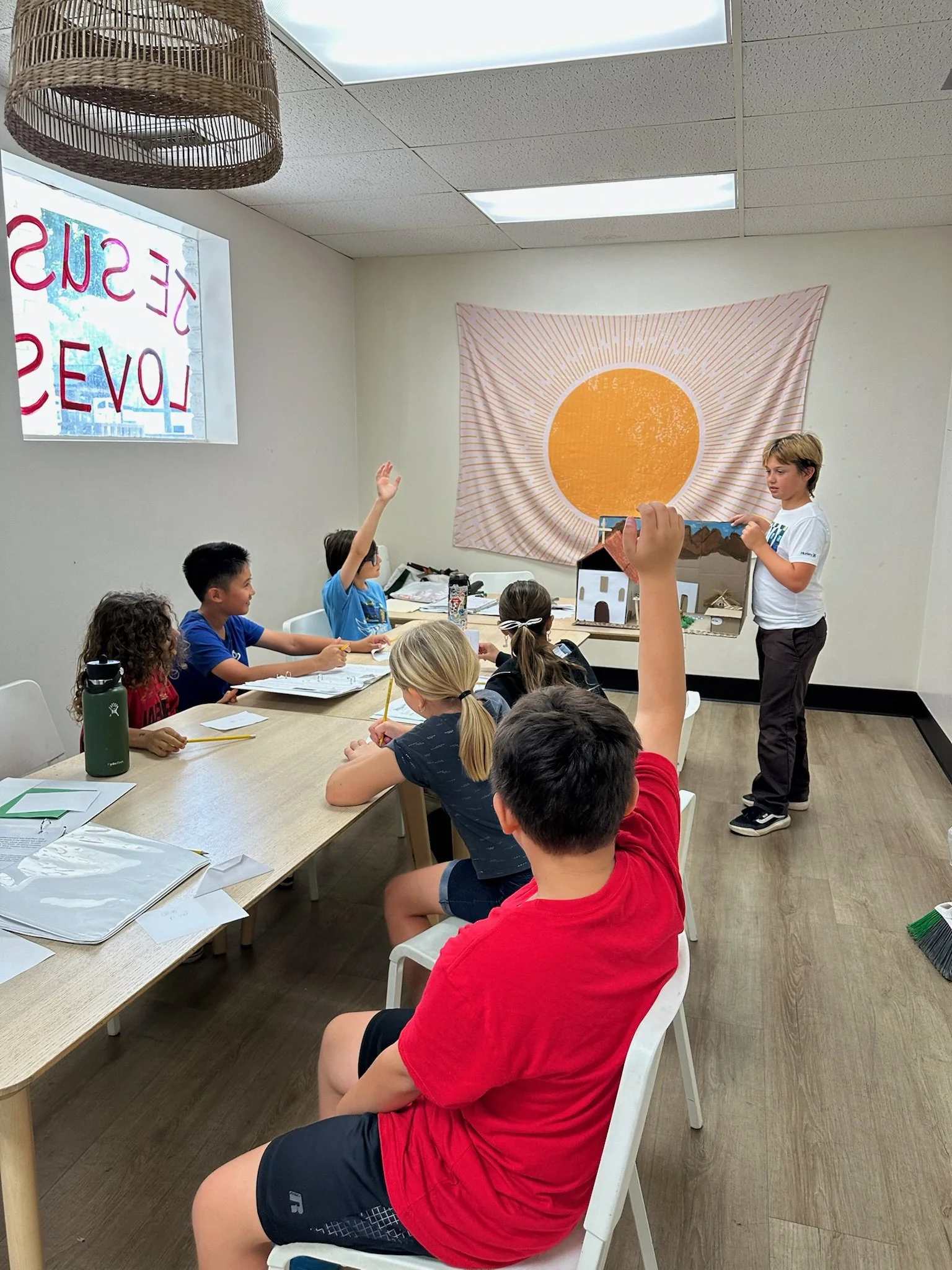 Indoor classroom scene with children seated at a long table, one child raising his hand and another standing at the front with a puppet house. A large orange sun tapestry decorates the wall, with fluorescent ceiling lights.