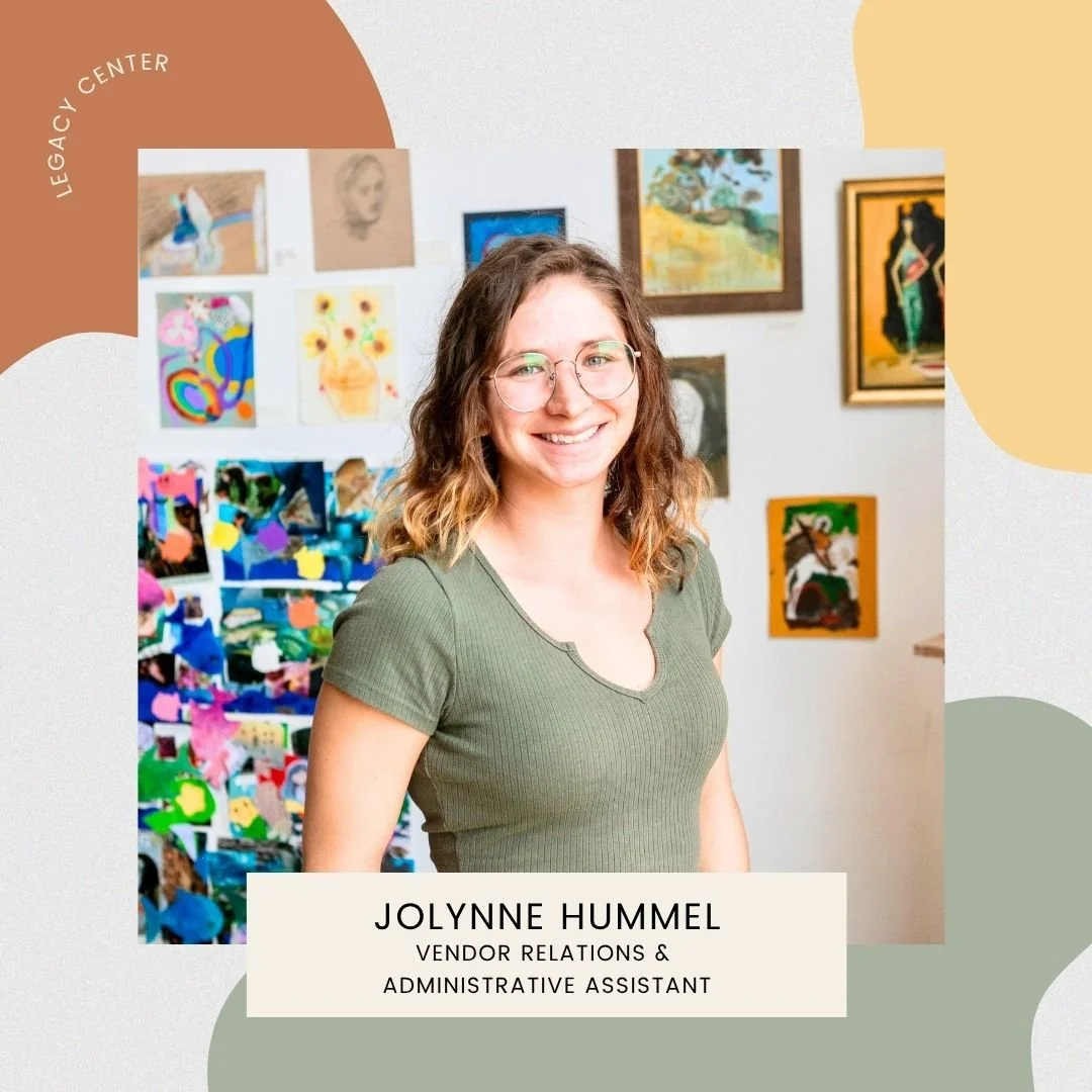 A young woman with glasses and wavy brown hair smiling at the camera, standing in front of a colorful art display on a gallery wall.
