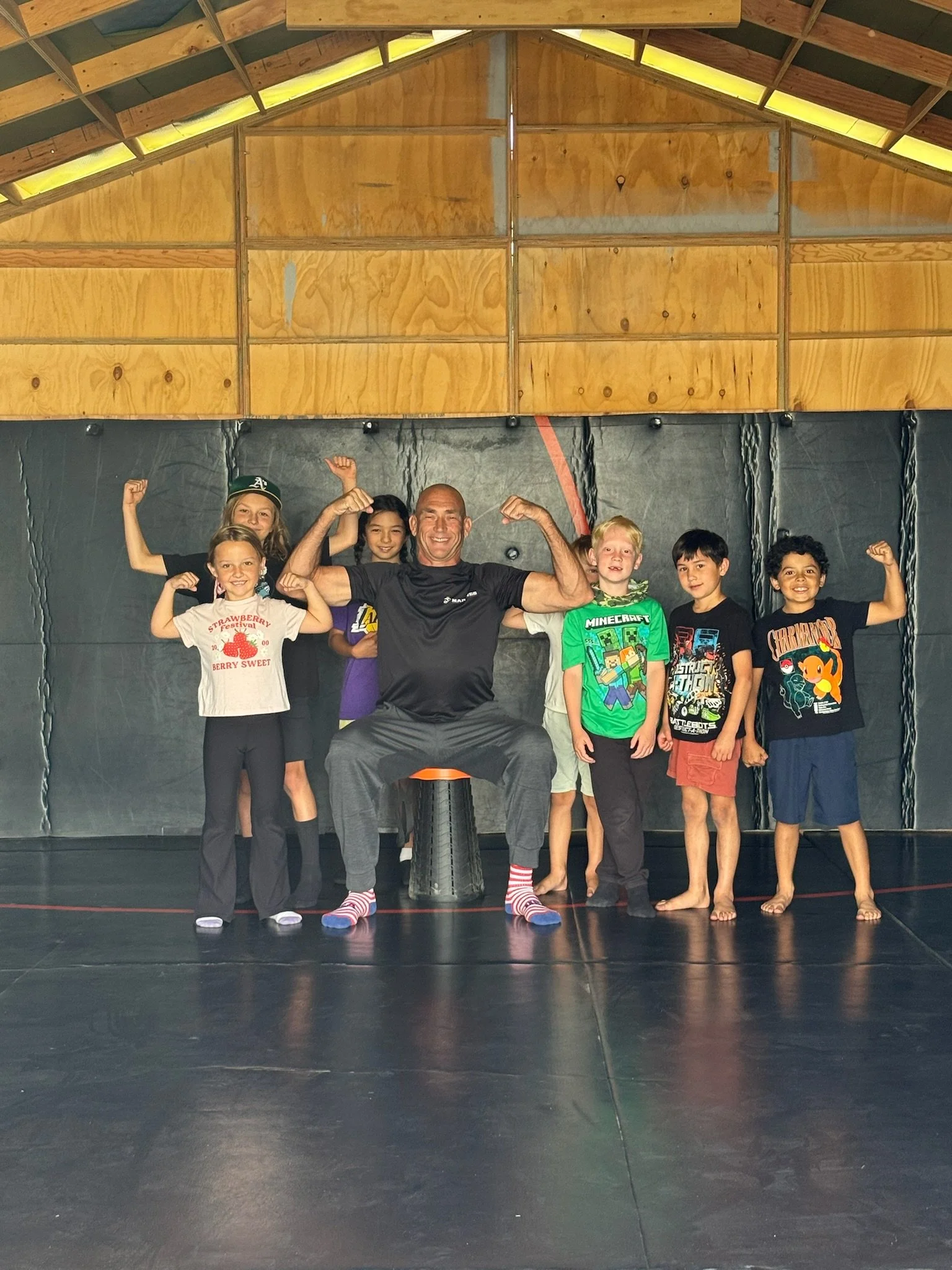 Group of six children and one adult, all smiling and flexing muscles, standing in a gymnasium with a black padded wall and wooden ceiling.
