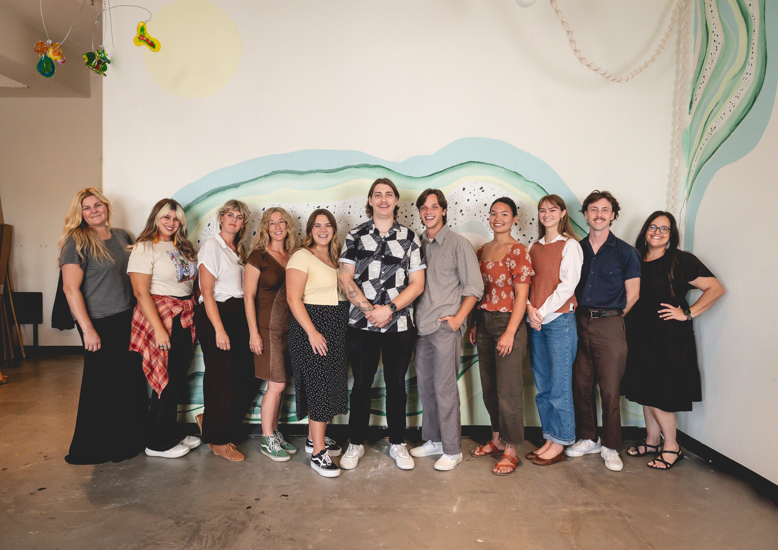 A group of thirteen diverse people standing side by side in front of a colorful abstract mural painting on a white wall, smiling at the camera.