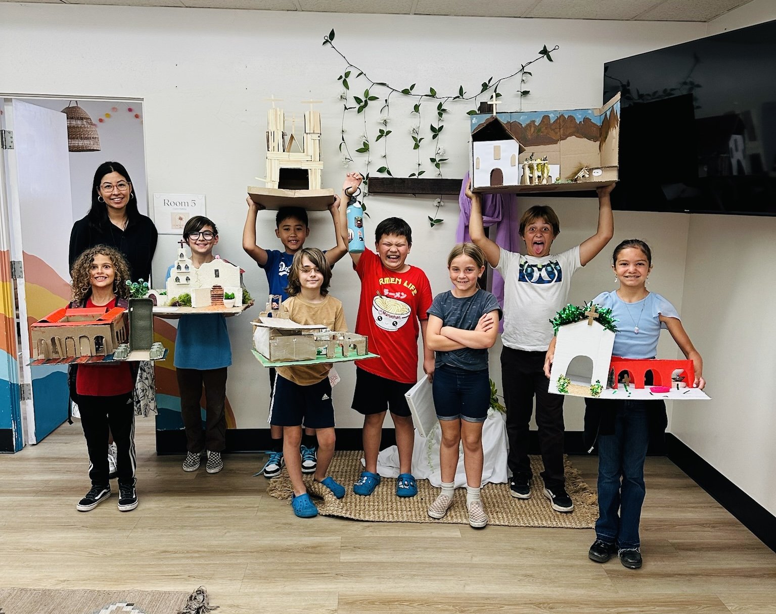 Group of children and a woman posing with their handmade miniature models of buildings, standing on a wooden floor in a room with white walls and a TV. The children are smiling and holding their models, which include churches and other structures, with some raising their models in the air.