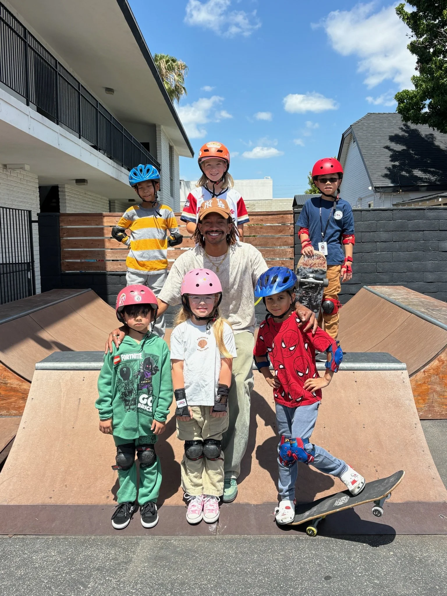 A group of children and a man are at a skatepark. The children are wearing helmets and protective gear. One of the boys is on a skateboard, and the man is standing behind some skateboard ramps, smiling.