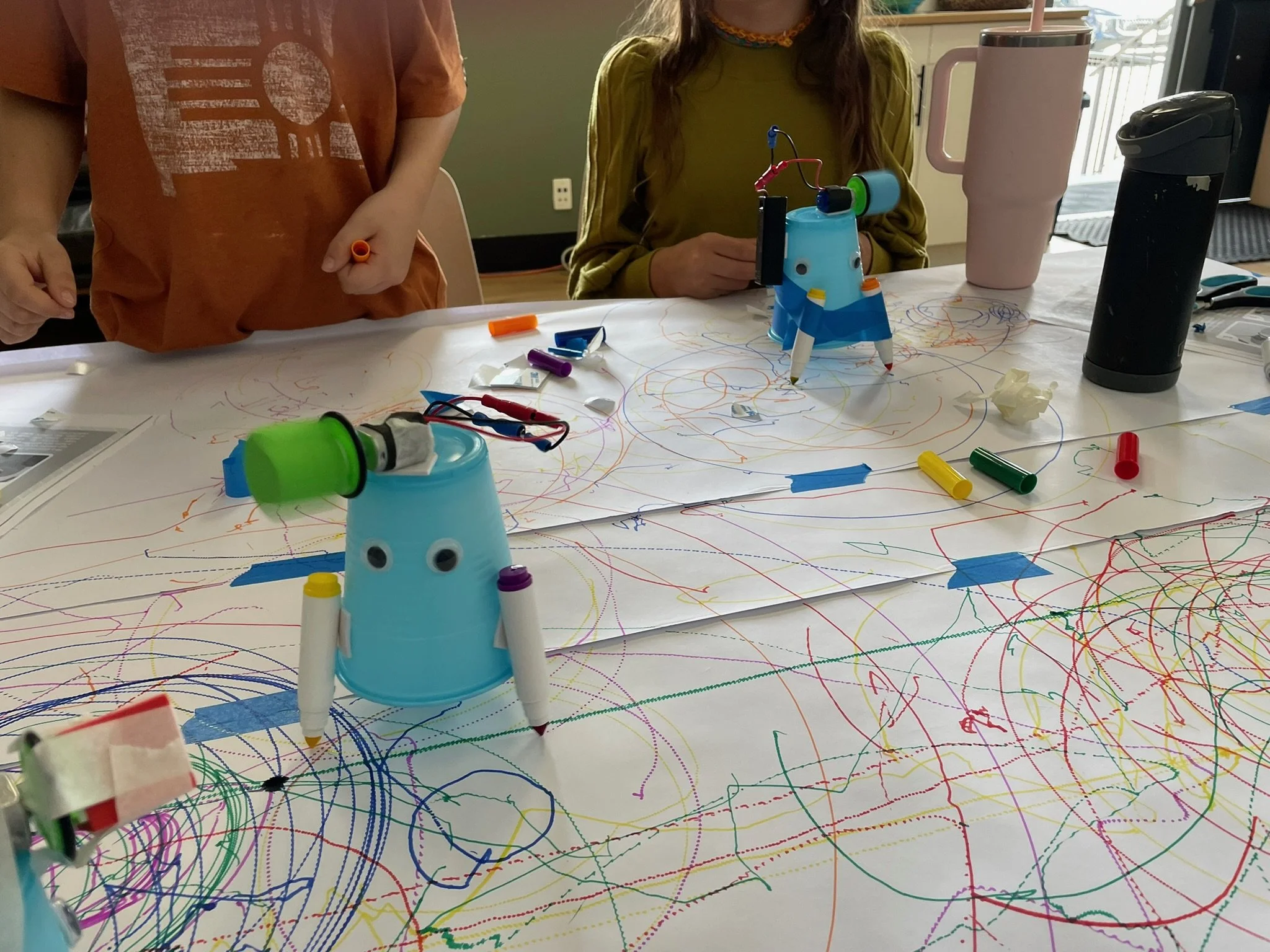 Children's artwork featuring a colorful, chaotic scribble drawing on a white paper with various markers, a small robot toy with googly eyes, a black thermos, a pink tumbler, and a black coffee cup on a table.