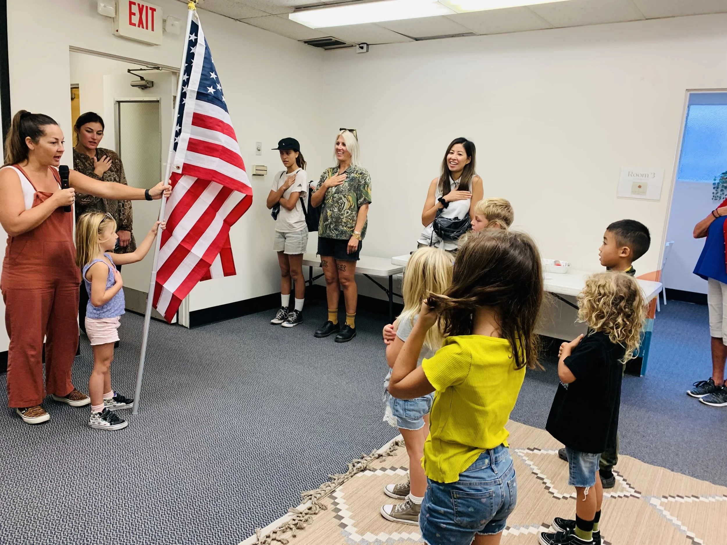 A group of children and adults standing inside a room with their right hands over their hearts, participating in a pledge or patriotic ceremony. One woman is holding an American flag. The room has a white wall, an exit sign, a mirror, and a window with a sign labeled 'Room 3'.