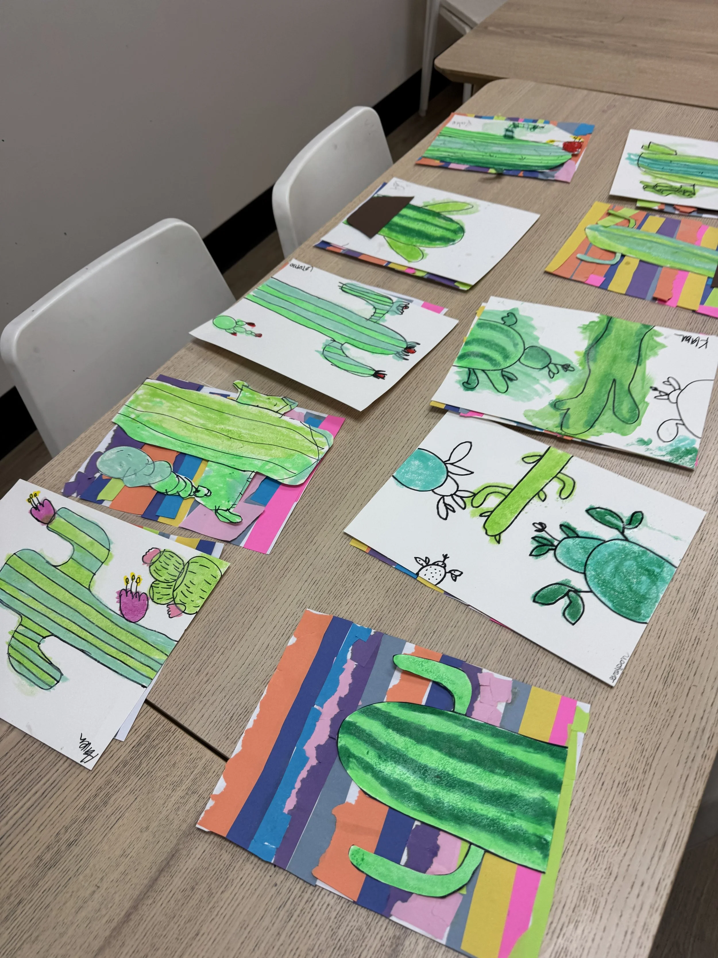 Children's artwork featuring various cactus illustrations, some colored green, on colorful paper backgrounds spread out on a wooden table.