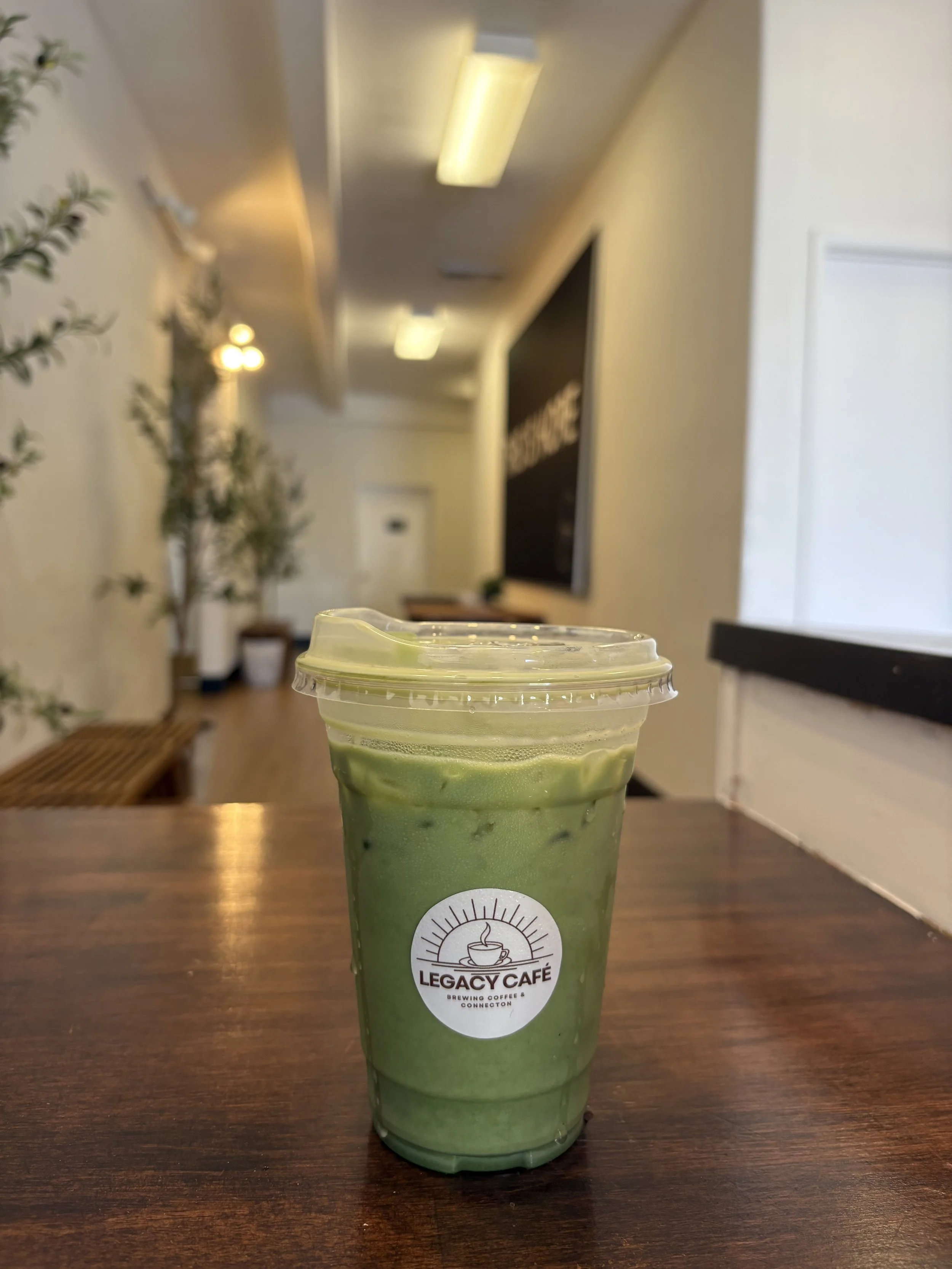 A green matcha latte in a clear plastic cup with a plastic lid, placed on a wooden table inside a coffee shop named Legacy Cafe.