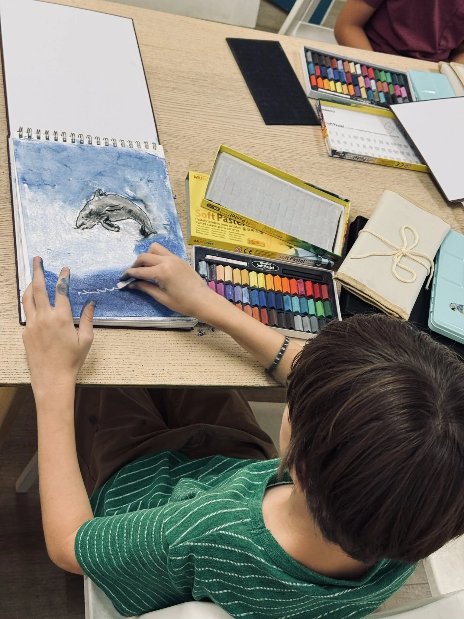 A boy with short brown hair wearing a green striped shirt is sitting at a desk, drawing in a sketchbook with a white pastel.
