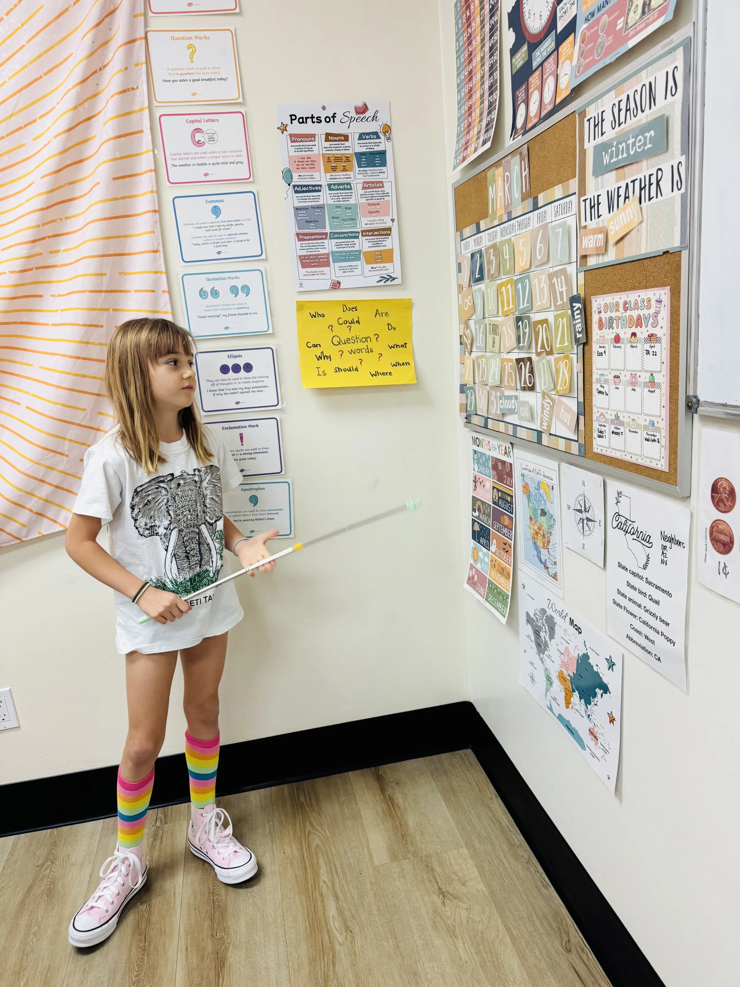 A young girl with rainbow socks and pink shoes stands in a classroom next to a white wall decorated with educational posters, a calendar, and maps, holding a pointer and looking to the side.