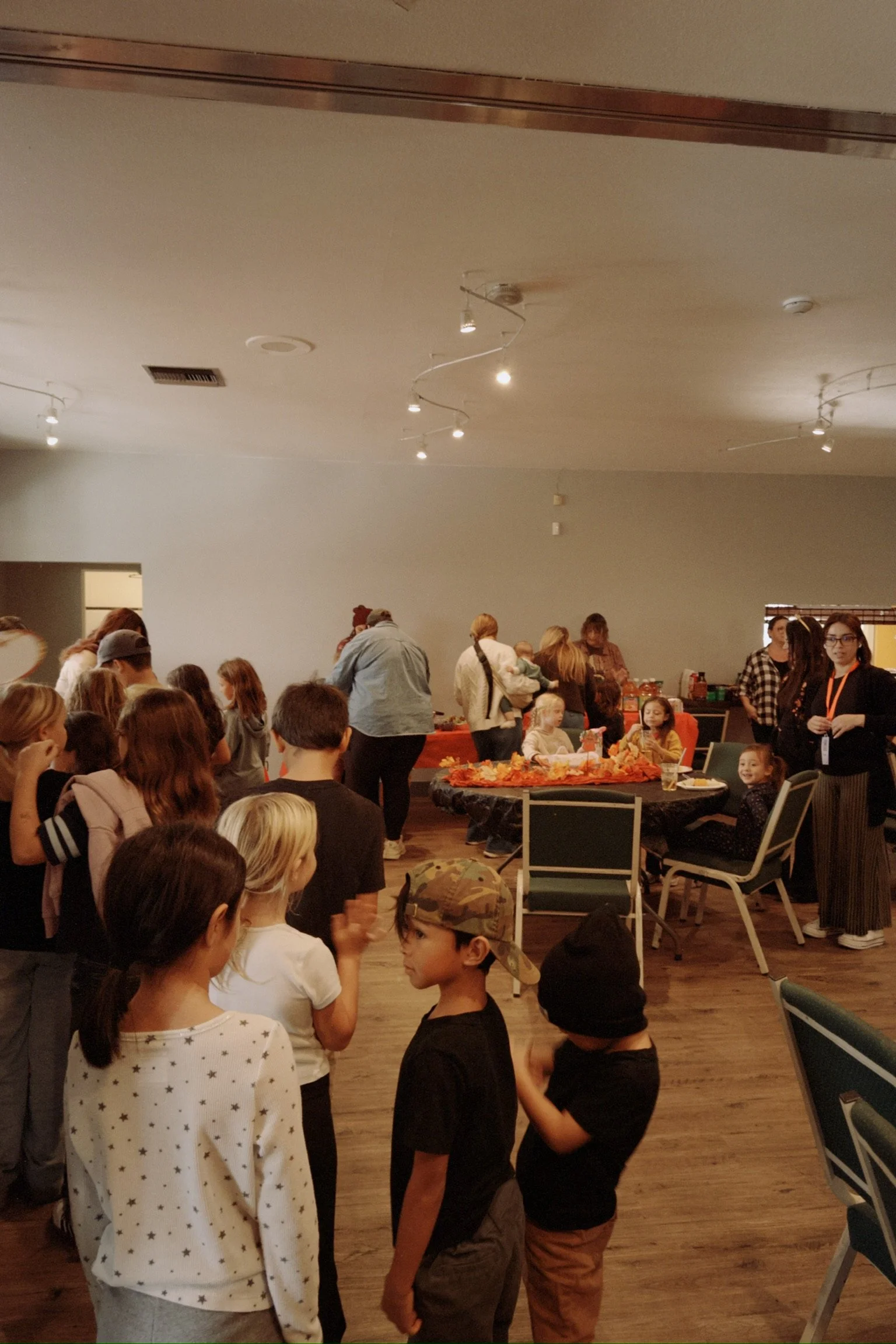 A group of children and adults gathering in a room with tables decorated with fall themed decorations, surrounded by chairs, in a social event or party setting.