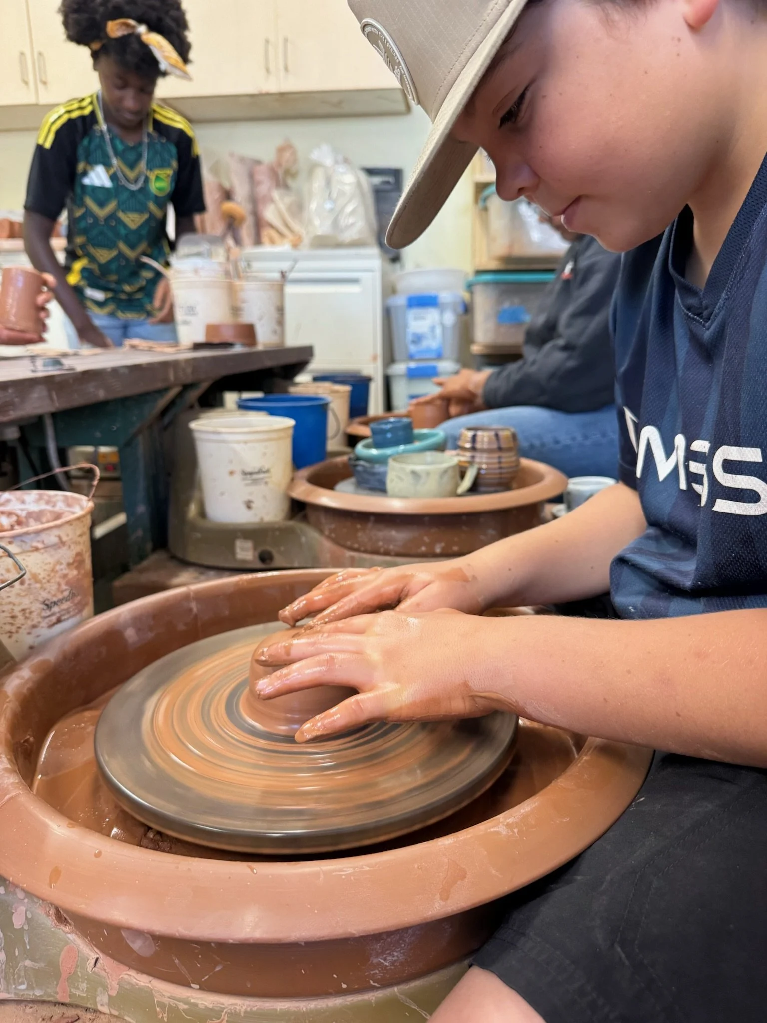 A person shaping clay on a spinning pottery wheel in a pottery studio, with other people working on similar projects in the background.
