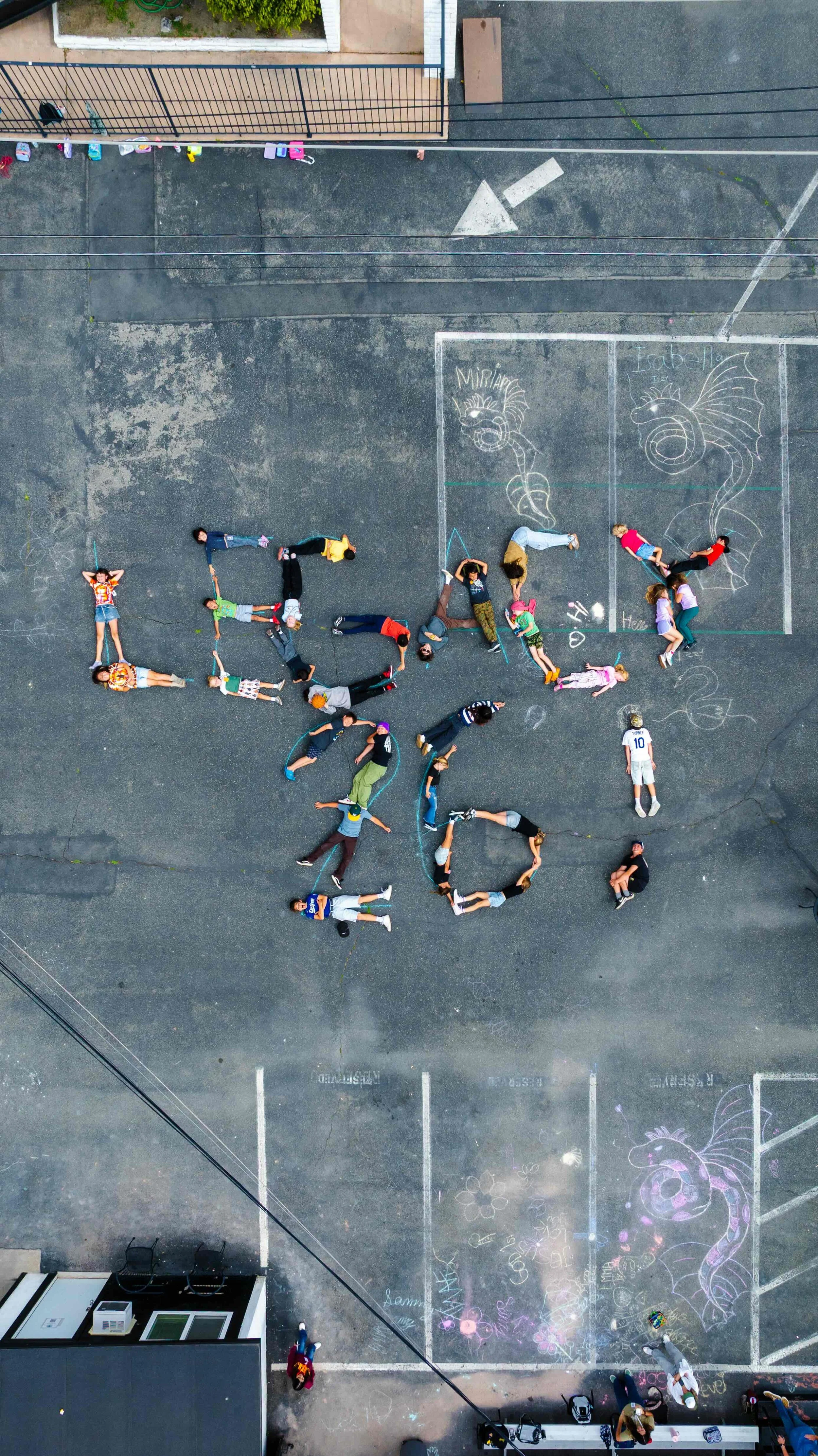 A group of children and adults lying on the ground forming the shape of the word 'LOVE'. There is chalk artwork on the ground including a dragon and various drawings.