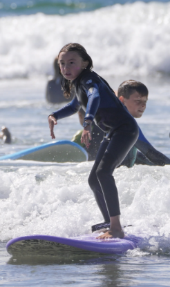 A girl standing on a surfboard in the ocean with waves, wearing a wetsuit, with a boy in the background also on a surfboard.
