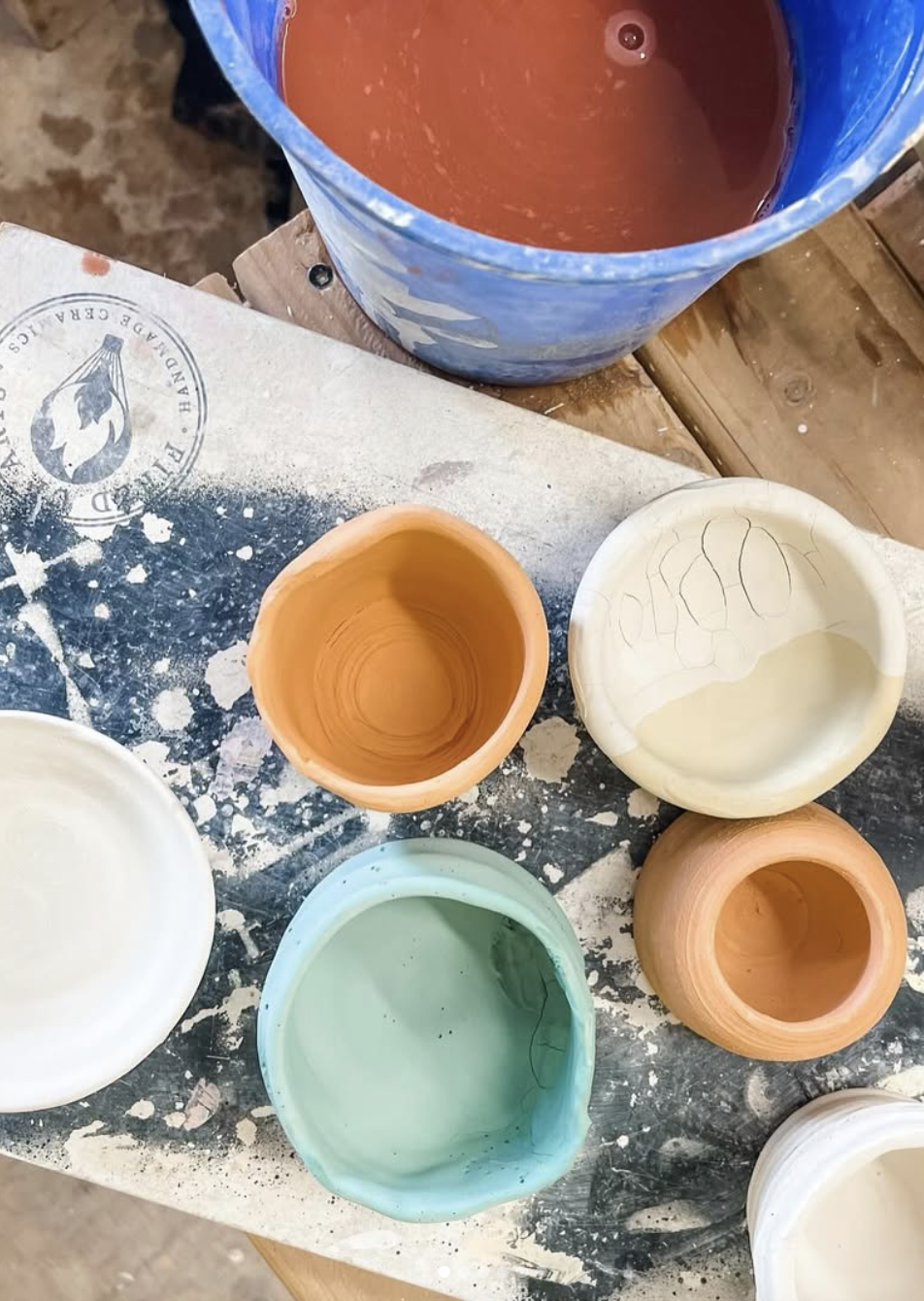 Colorful ceramic cups and a bucket of red paint on a work table covered with splatters of paint.