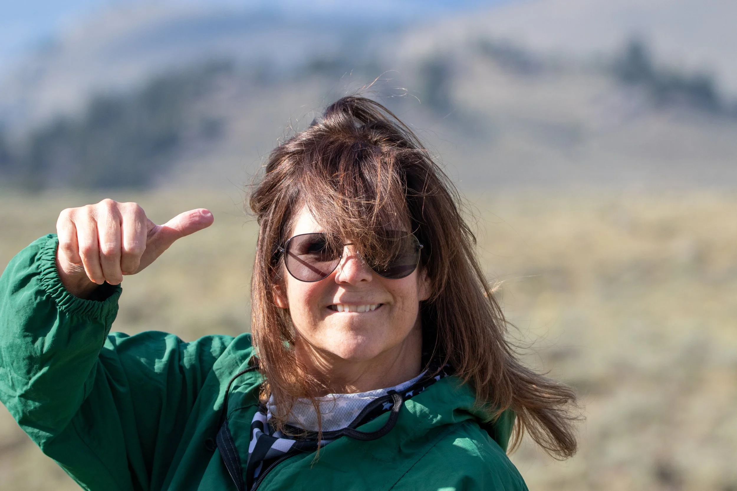 A woman with brown hair wearing sunglasses, a green jacket, and a patterned neck gaiter, smiling and pointing with her thumb at her head in an outdoor setting.