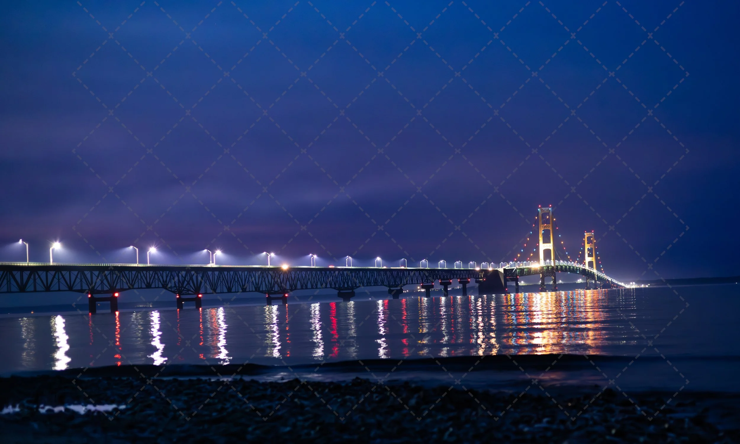 A brightly lit bridge over calm water at night, with reflections visible on the surface and a dark, cloudy sky above.