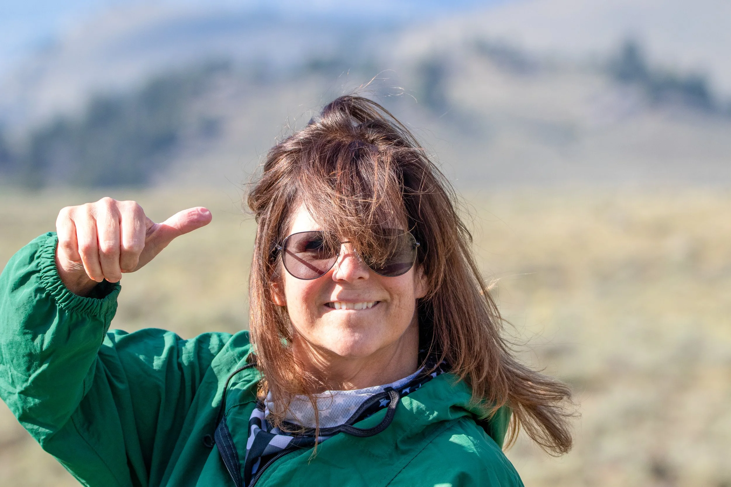 A woman with long brown hair wearing sunglasses and a green jacket, smiling and giving a thumbs-up outdoors with a blurred landscape in the background.