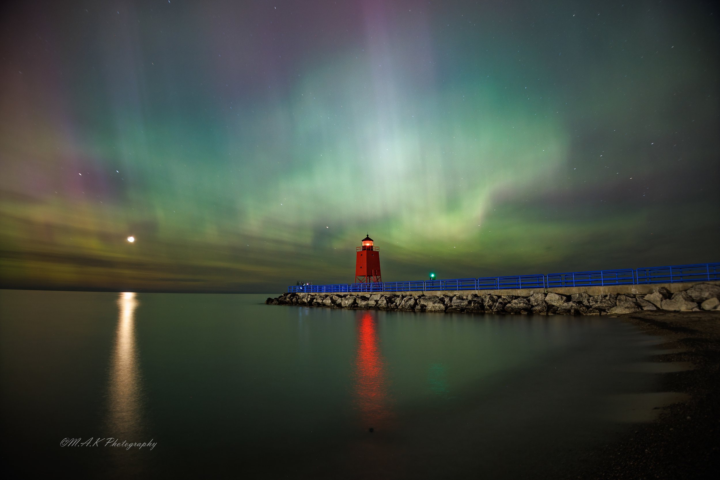 Nighttime scene of aurora borealis over a lighthouse on a rocky pier, with reflections on calm water.