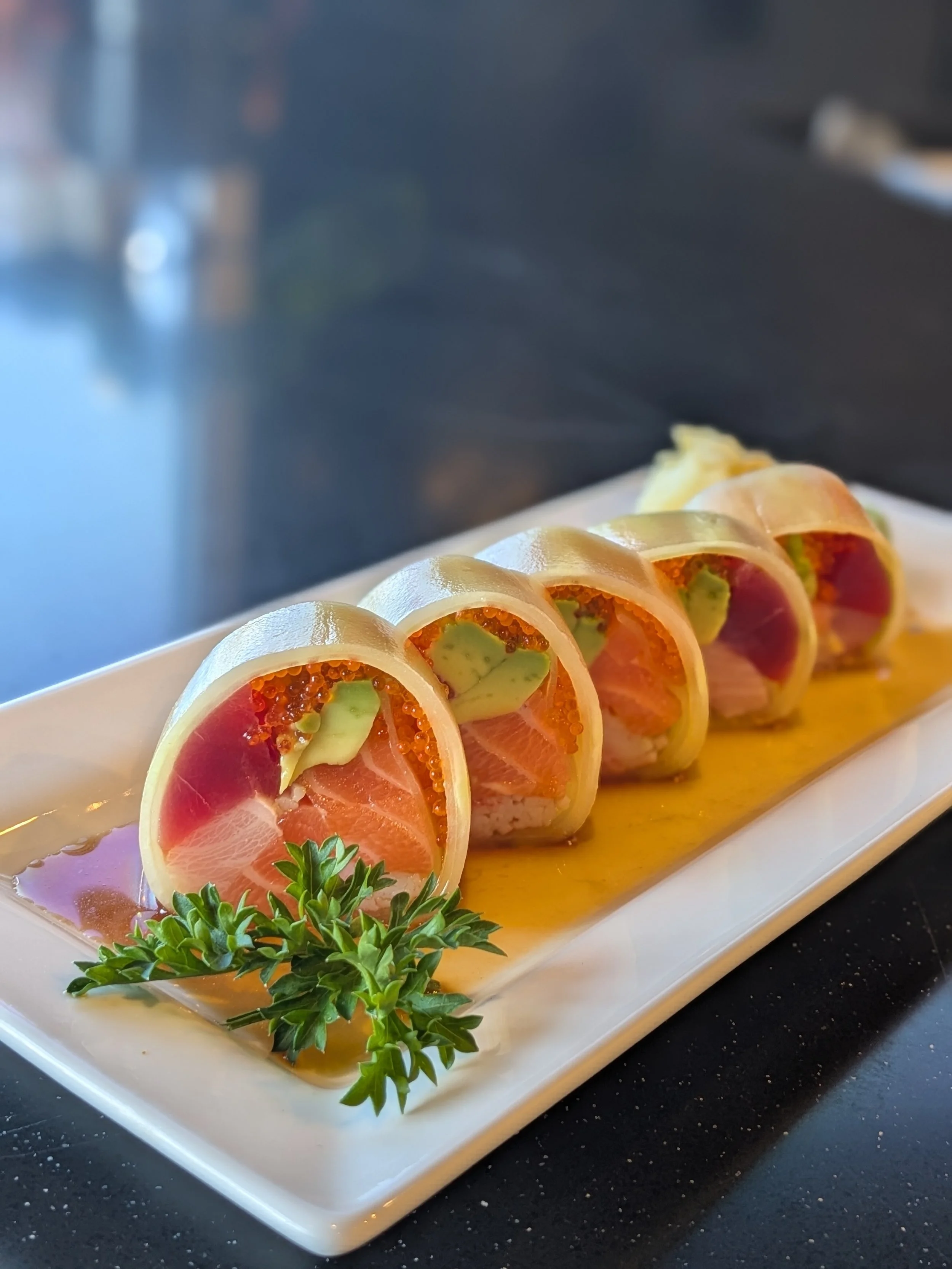 A plate of sushi rolls filled with salmon, avocado, and orange fish roe, garnished with parsley, on a white rectangular dish with a yellow sauce underneath.