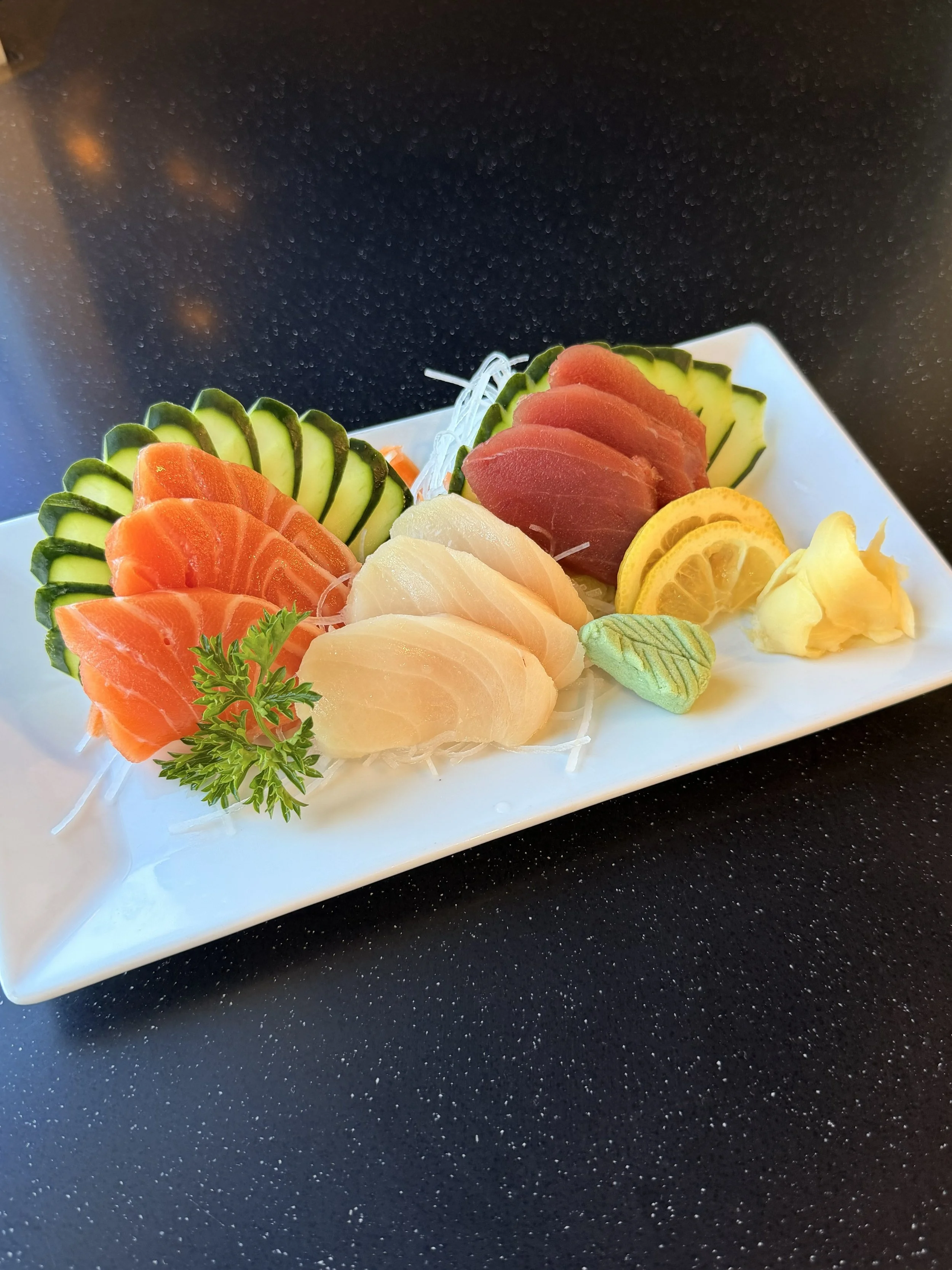 Assorted sashimi slices of salmon, tuna, and white fish on a white rectangular plate with cucumber, lemon, and wasabi garnishes, on a black speckled surface.