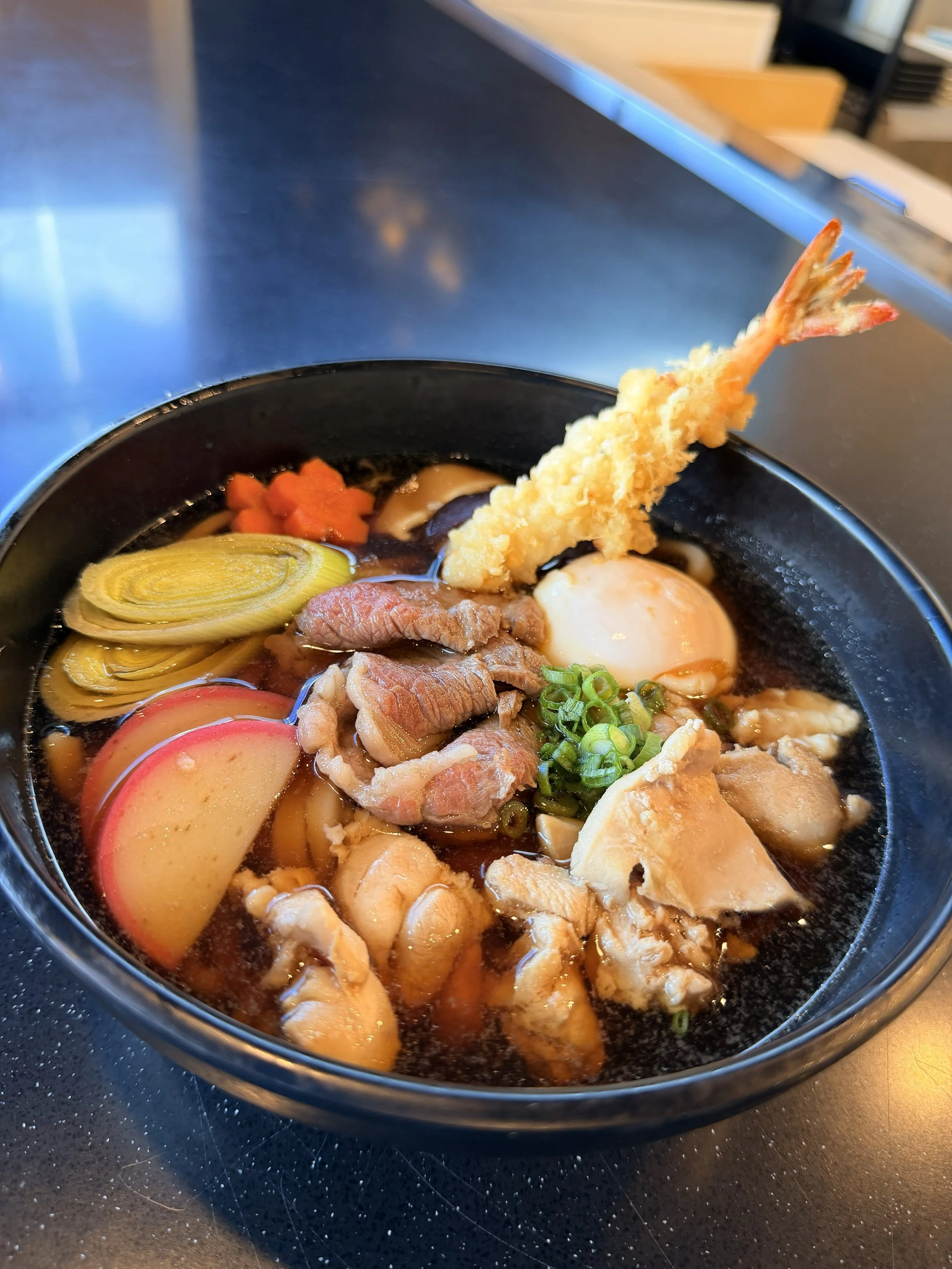 A bowl of Japanese ramen topped with a tempura shrimp, sliced fish cakes, green onions, vegetables, and a soft-boiled egg.