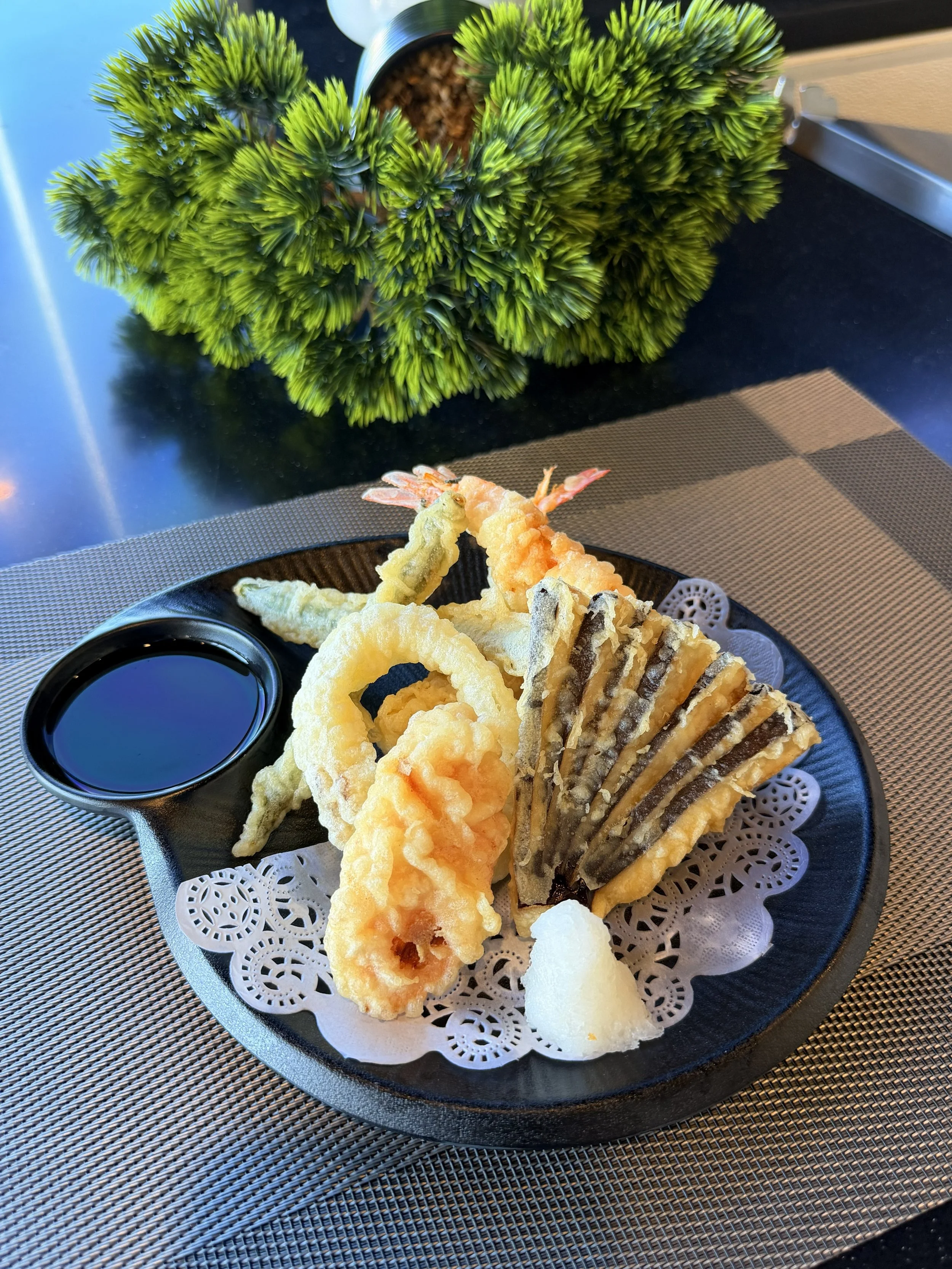 Plate of assorted tempura including shrimp, eggplant, kabocha squash, and onion rings with a small dish of soy sauce on the side. In the background, a potted succulent plant.