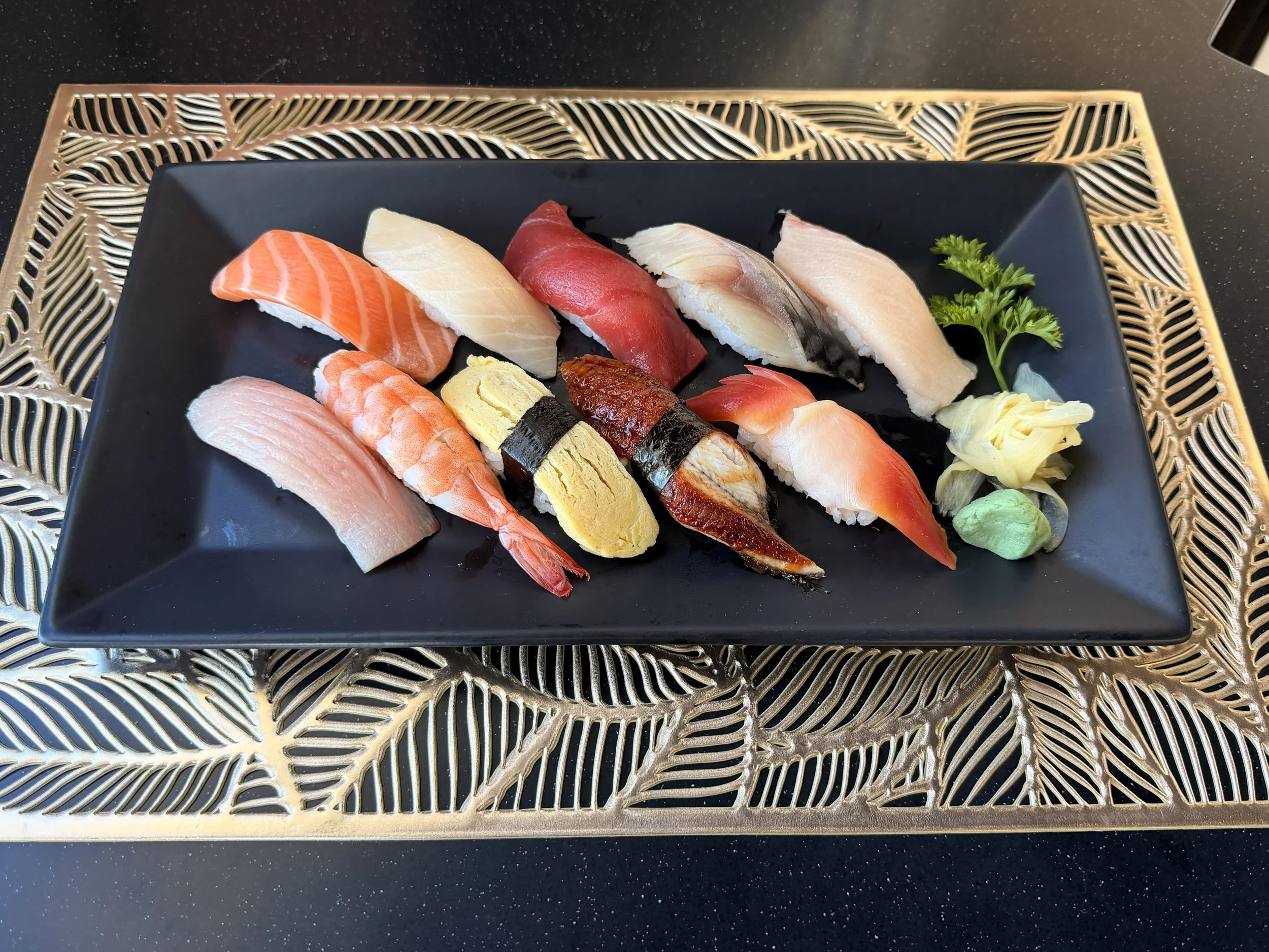 Assorted sushi pieces on a black plate with a garnish of parsley, on a black surface with a decorative gold and black patterned placemat.