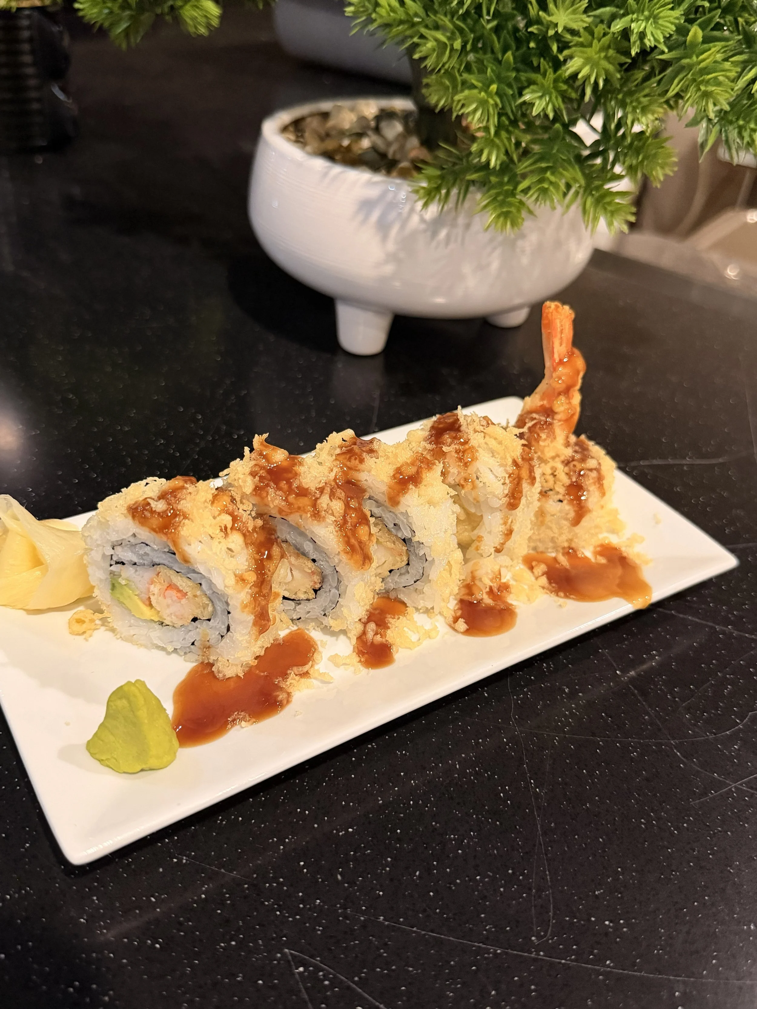Sushi rolls with tempura shrimp topped with sauce served with wasabi and pickled ginger on a white rectangular plate. In the background, a potted green plant is visible.