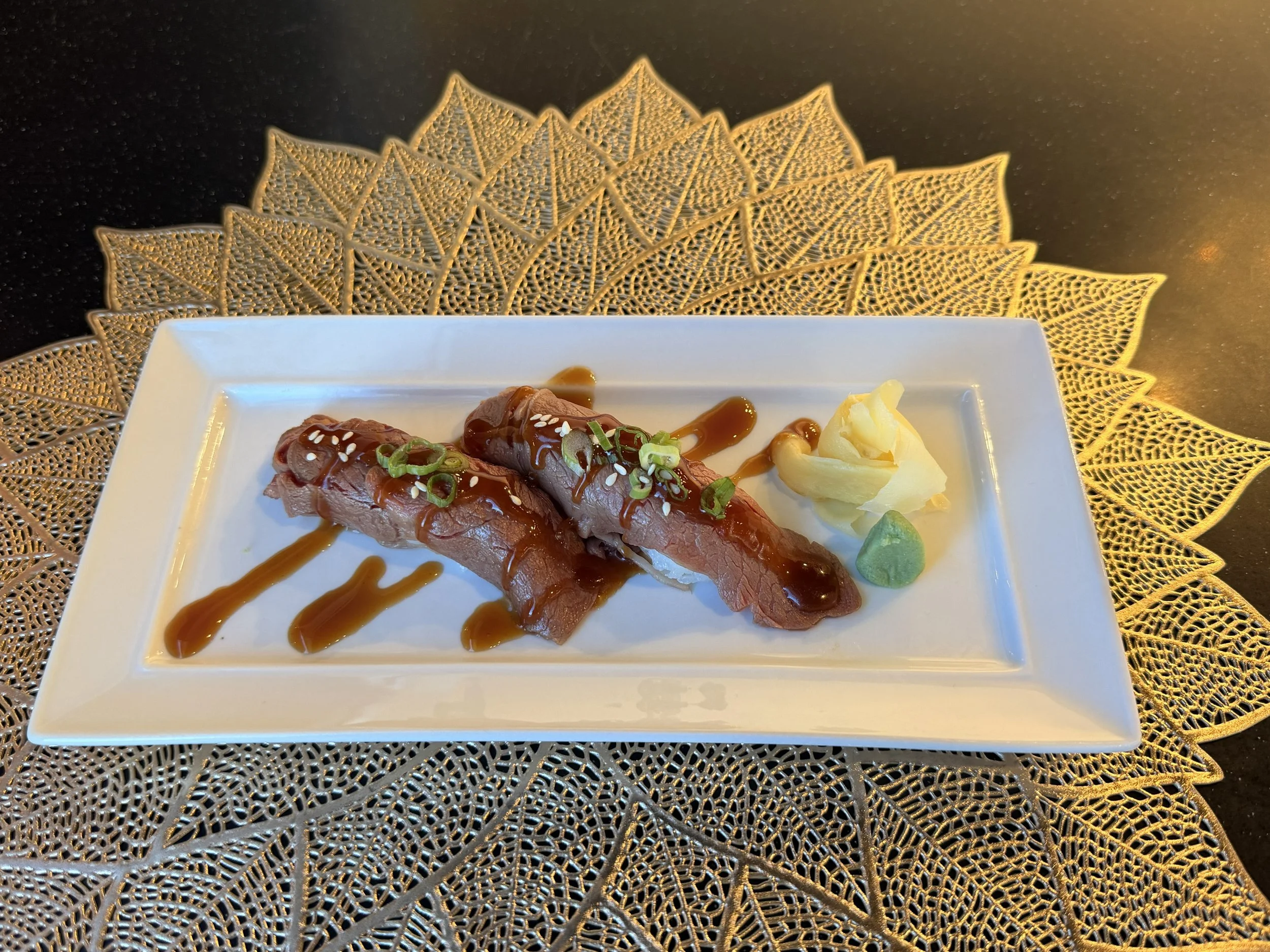 Sliced raw beef topped with green onions, sesame seeds, and sauce, served with wasabi and pickled ginger on a white rectangular plate, placed on a decorative gold lace placemat against a dark background.