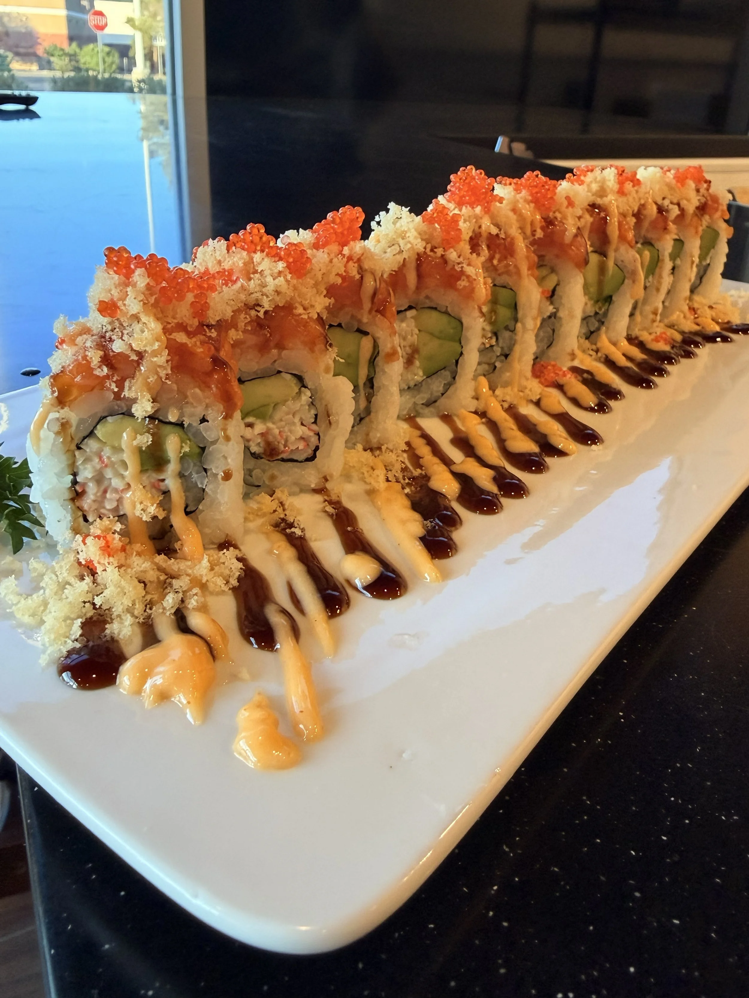 A plate of sushi rolls topped with orange fish roe, drizzled with various sauces, served on a white rectangular plate on a black countertop.