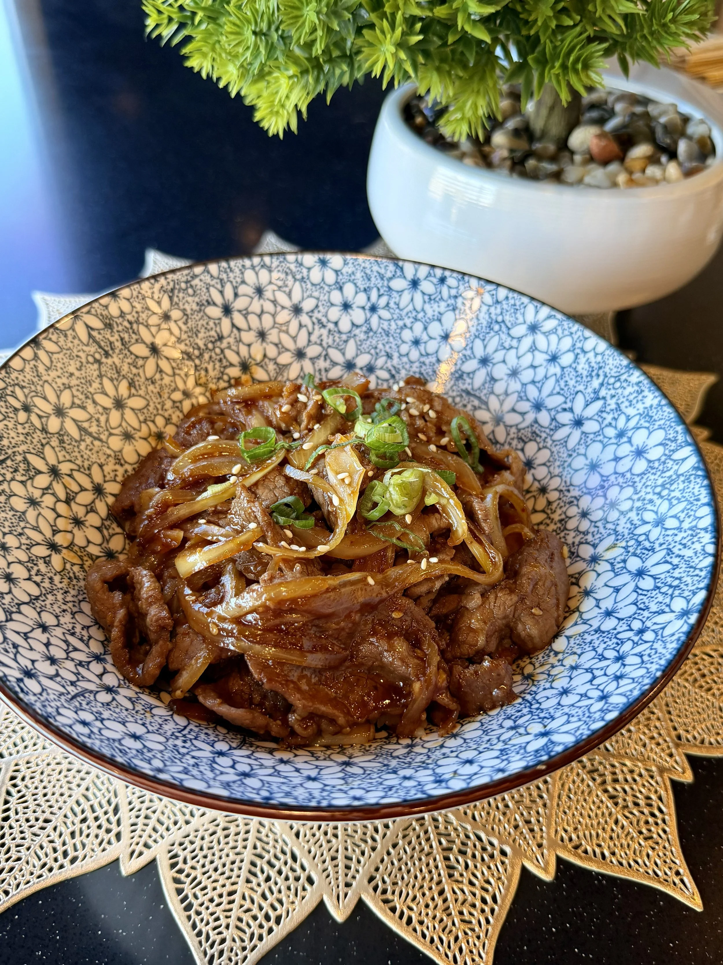 A blue and white floral patterned bowl filled with stir-fried beef and onions topped with chopped green onions and sesame seeds, placed on a decorative golden placemat.