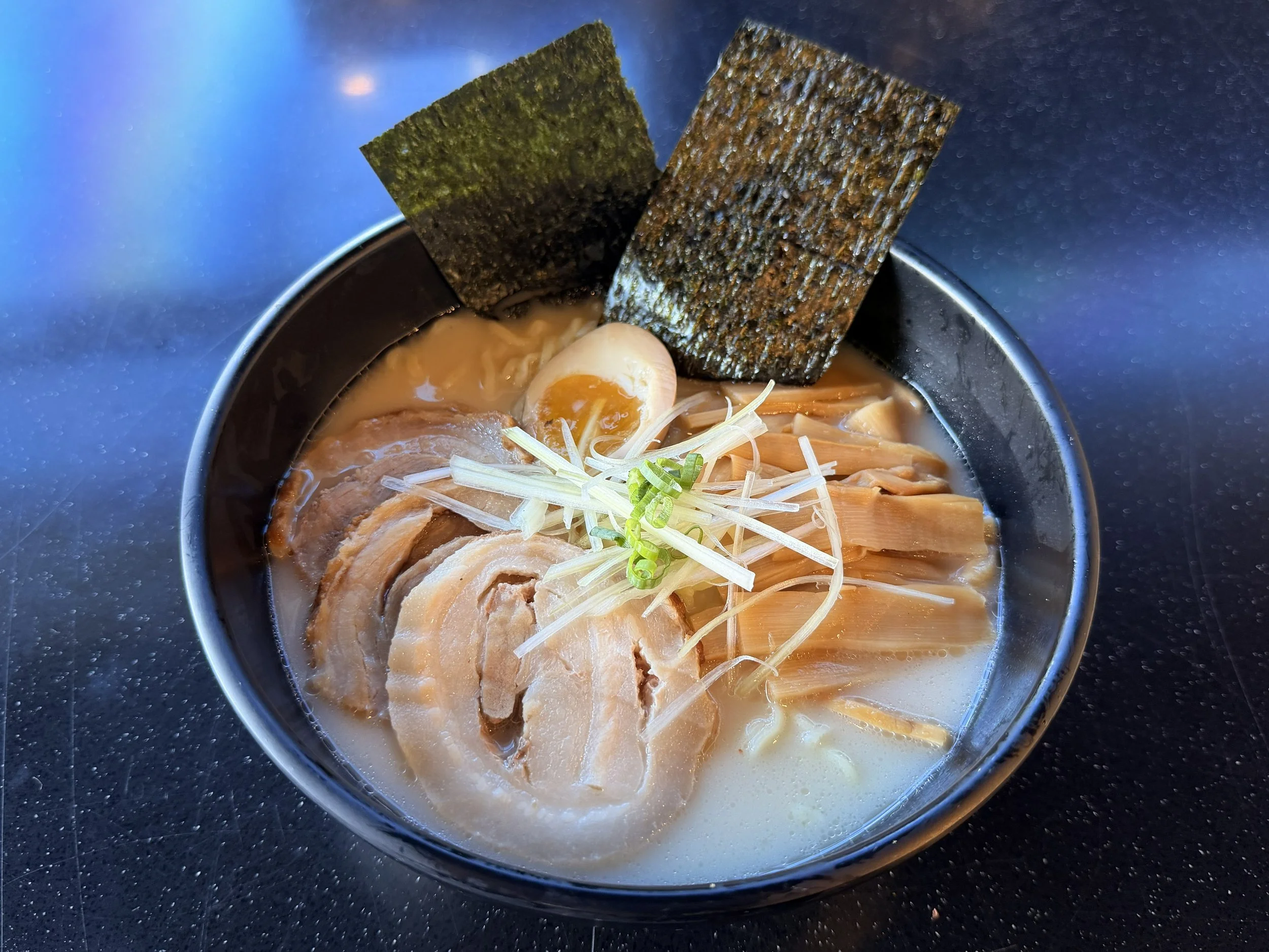 A bowl of ramen noodles with slices of pork, half a soft-boiled egg, shredded green onion, bean sprouts, and two sheets of nori seaweed on top.