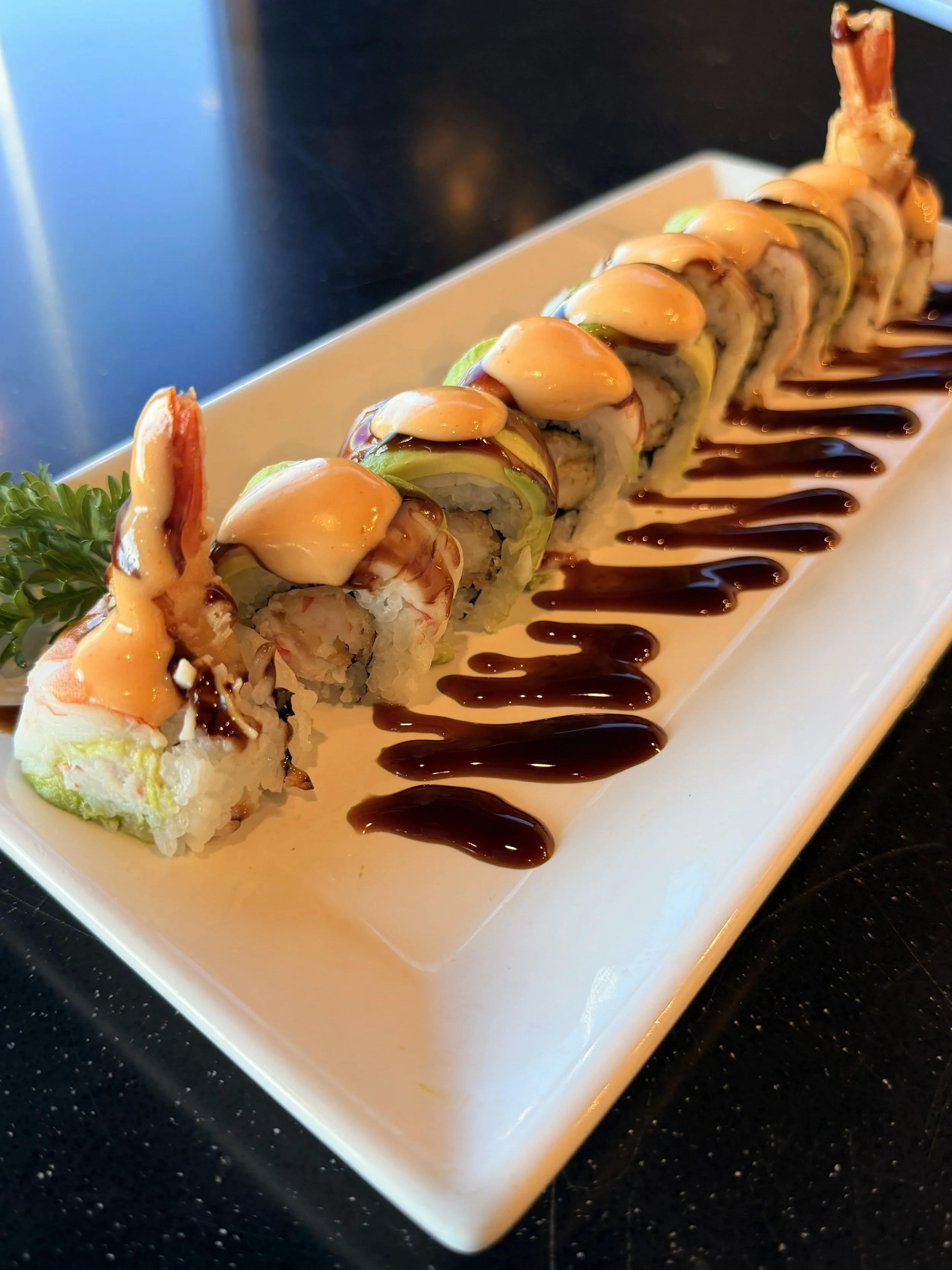 A plate of sushi rolls drizzled with eel sauce and topped with spicy mayo, garnished with a sprig of parsley, on a rectangular white plate.