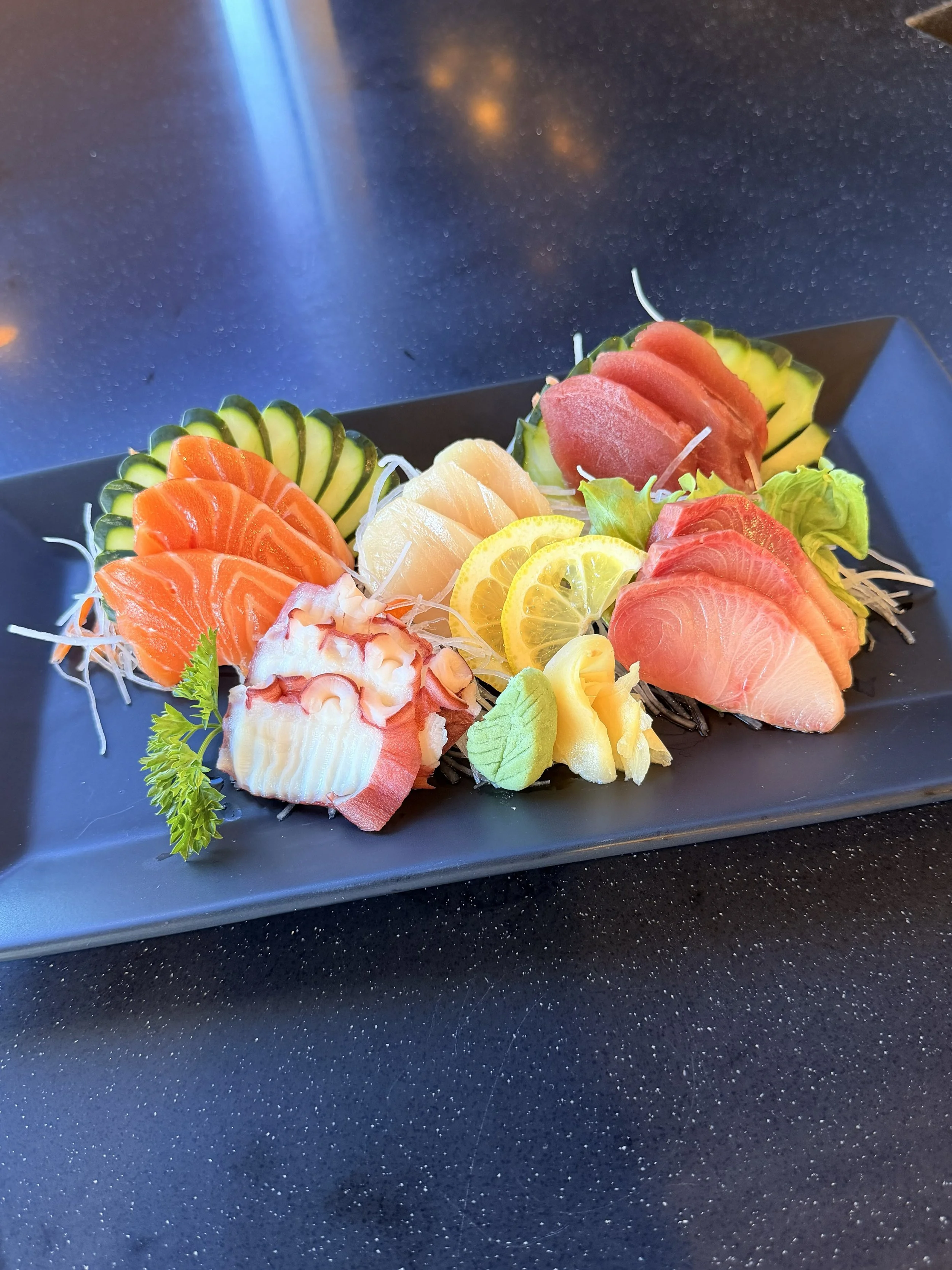 Assorted sashimi slices of salmon, tuna, and white fish, garnished with lemon slices, cucumber, and wasabi on a black rectangular plate.