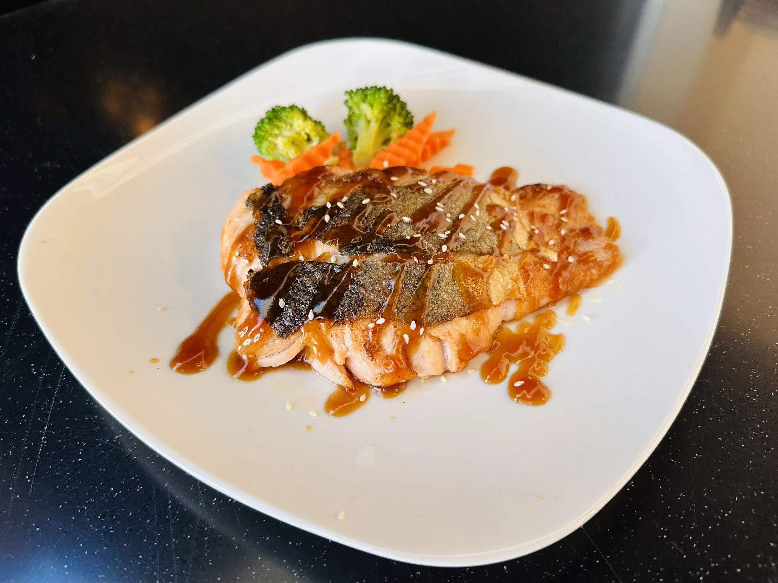 Plate with grilled salmon topped with sauce and sesame seeds, accompanied by broccoli, carrots, and spiralized carrots.